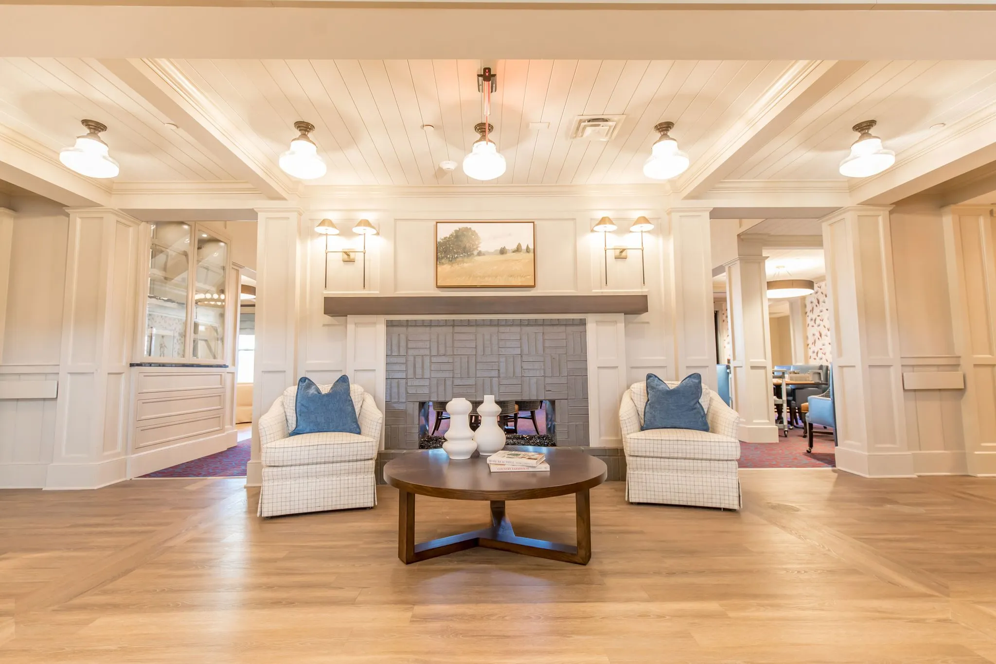 Senior living lobby with a tiled fireplace between schoolhouse pendants, cream plaid armchairs facing it