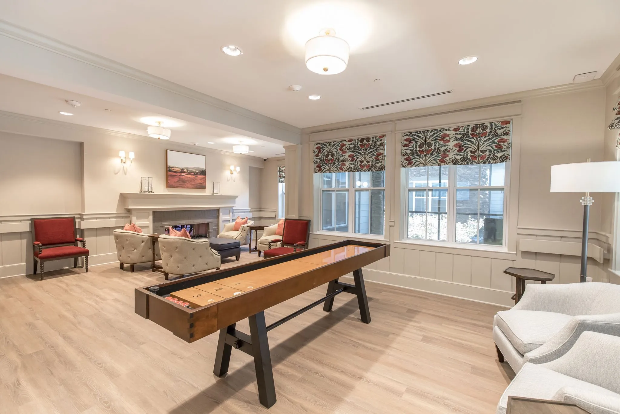 Senior living shuffleboard lounge with a full-length table on wood flooring, red chairs by the fireplace