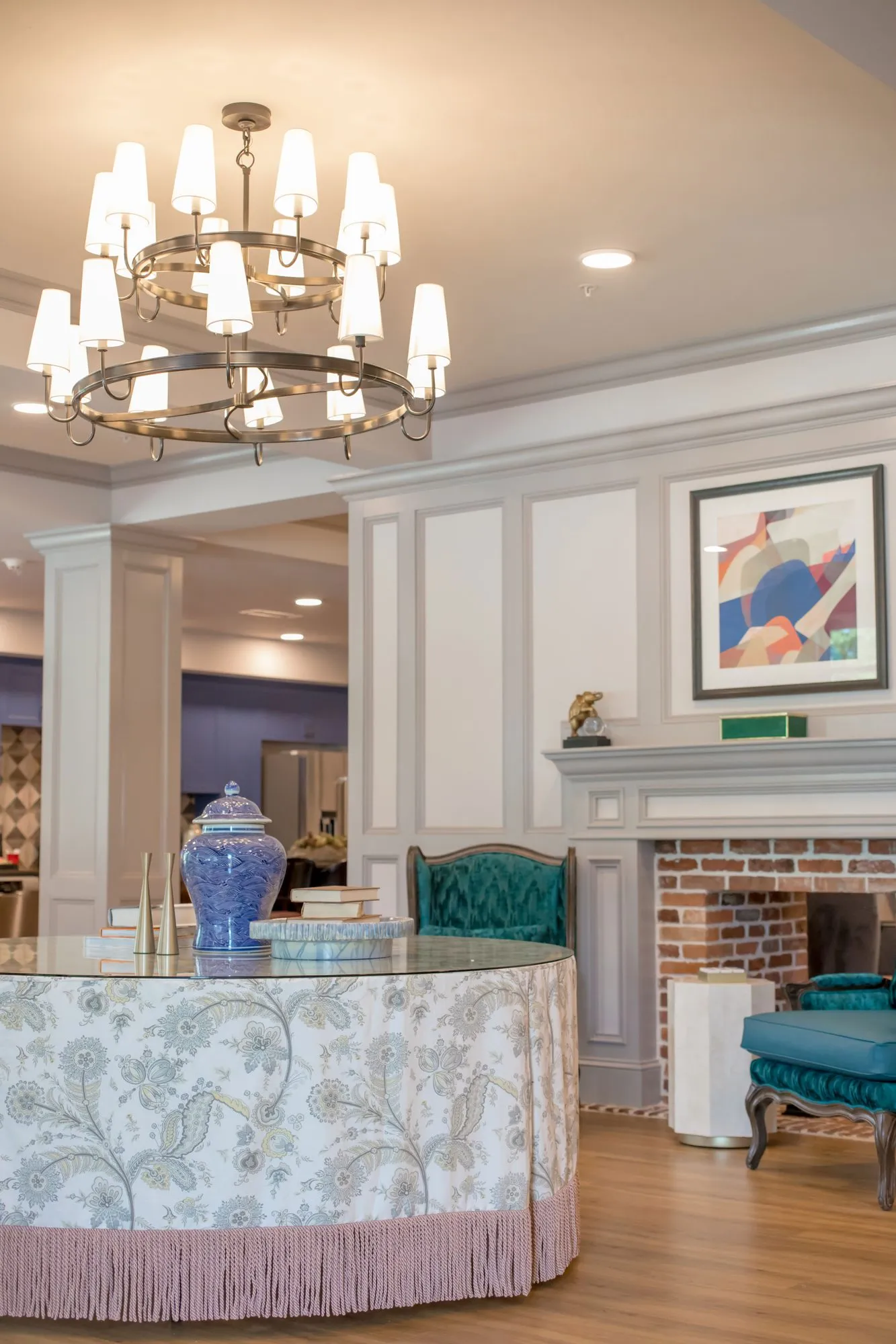 Senior living lobby with a round table skirted in paisley and pink fringe under a tiered candle chandelier, a brick fireplace