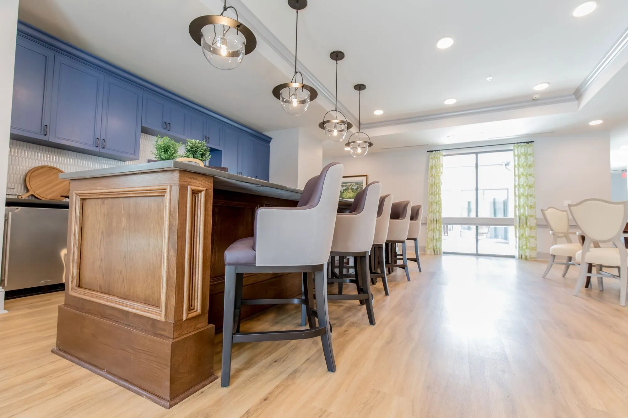 Senior living kitchen with a raised wood panel island and grey upholstered counter stools under glass globe pendants