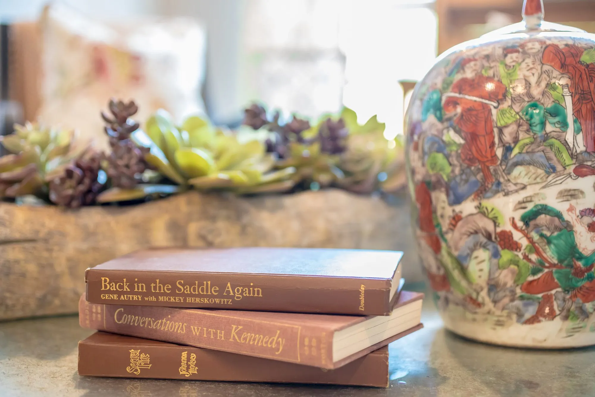 Senior living interior detail: old leather-bound books stacked beside a painted ginger jar and a wood bowl of succulents