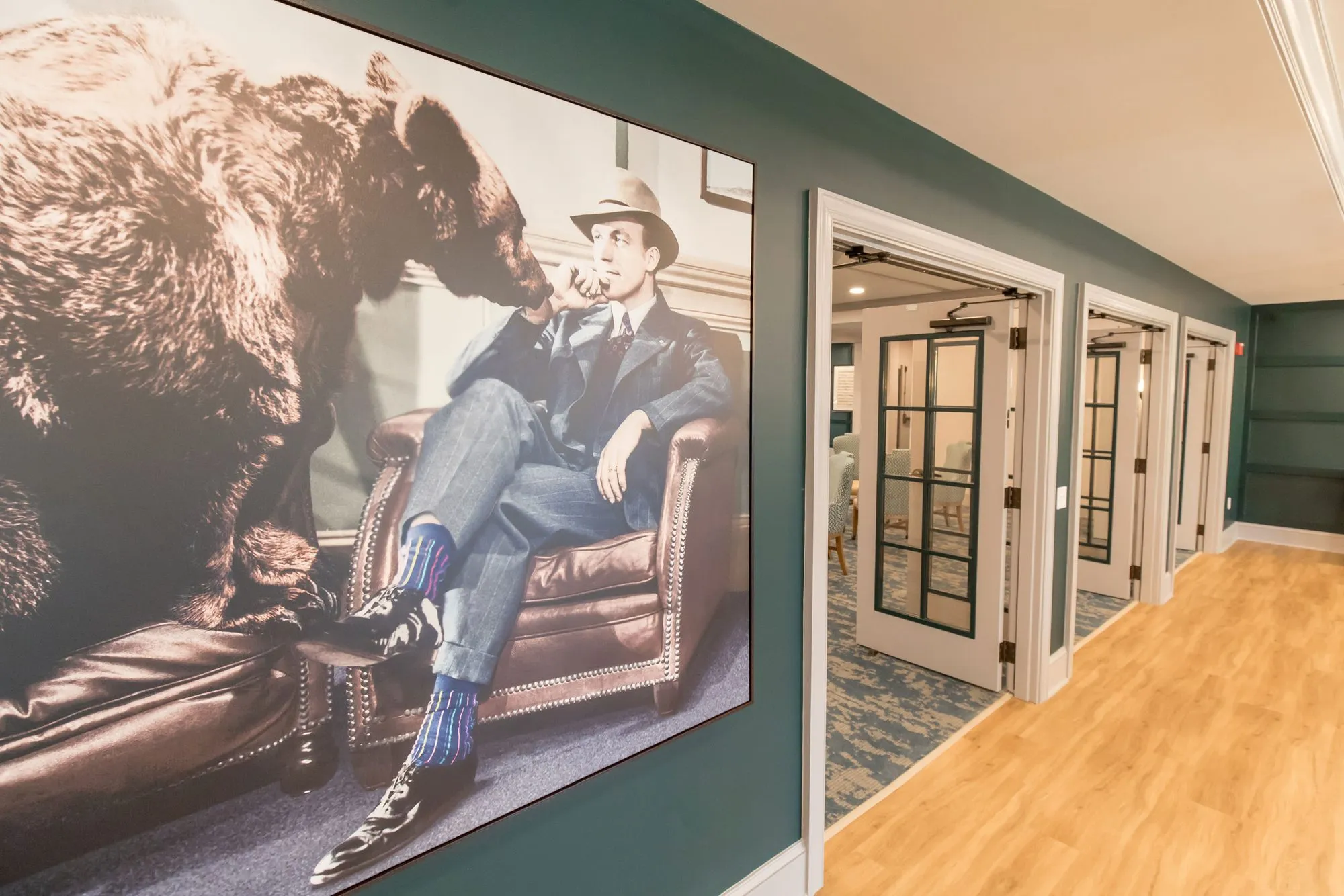 Senior living teal corridor hung with a large photograph of a man in a leather club chair beside a bear, glazed doors beyond