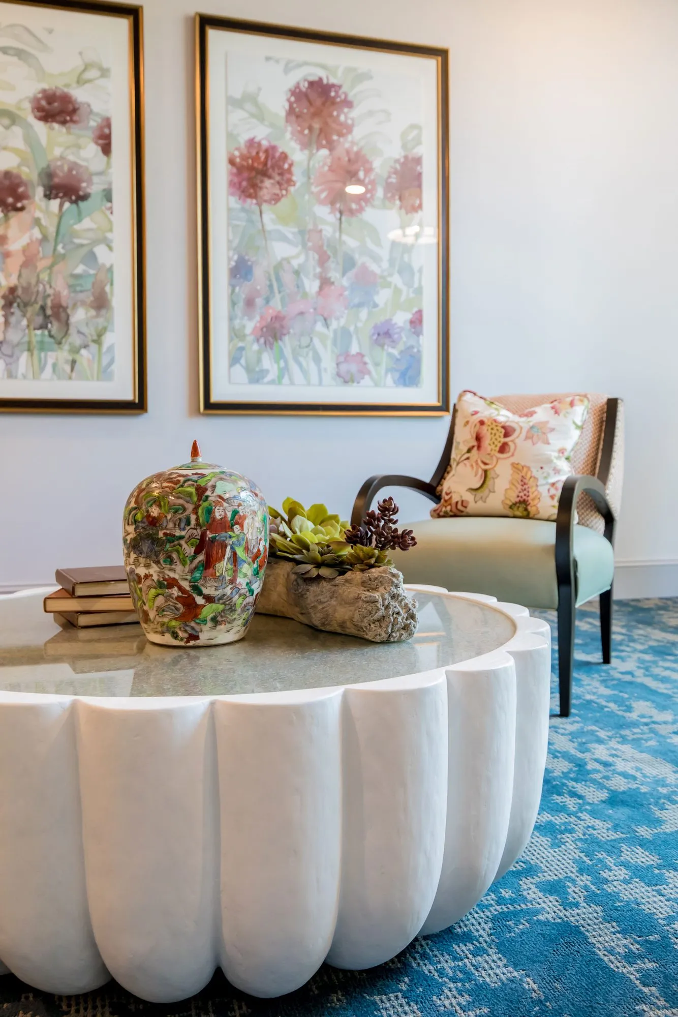 Senior living sitting room corner with floral watercolours above a patterned armchair and scalloped white table on a blue rug