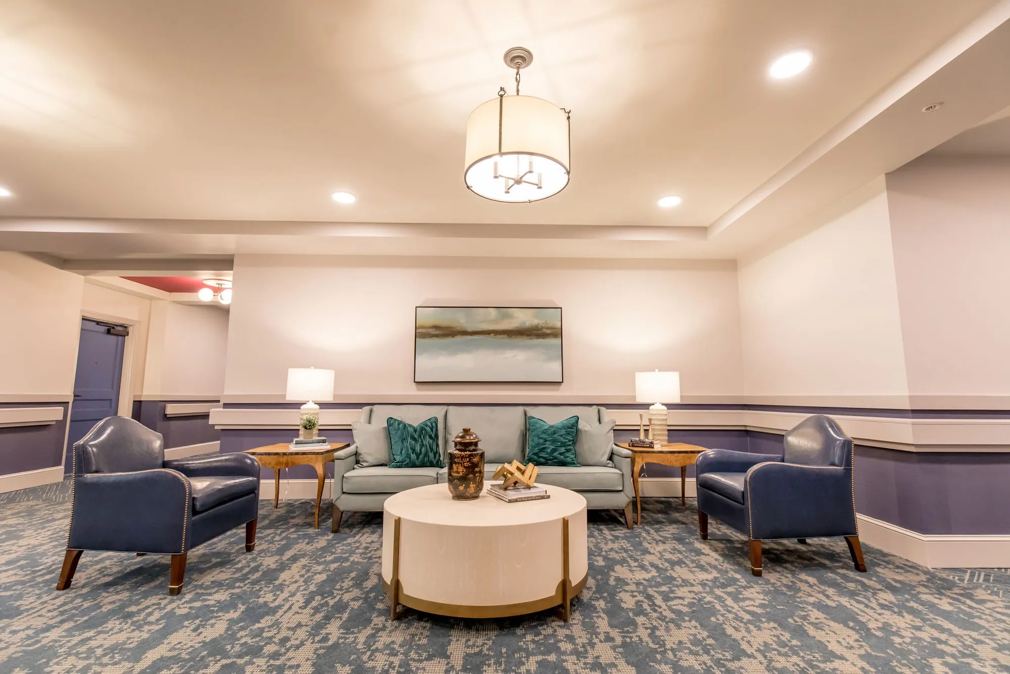 Senior living sitting area with a sage sofa between navy leather armchairs, a round white coffee table and a drum pendant