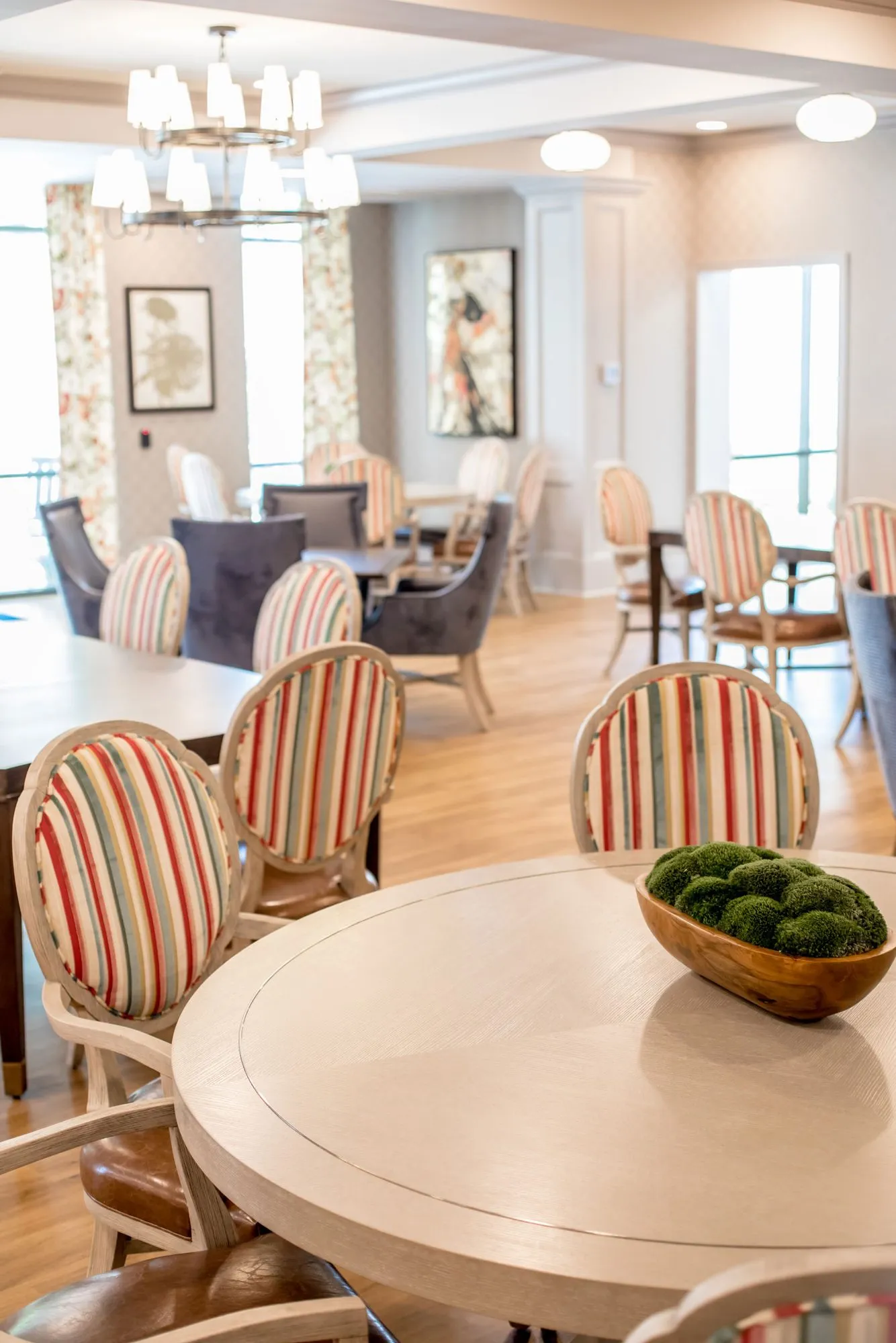 Senior living dining room with round tables and striped and navy chairs under a tiered chandelier