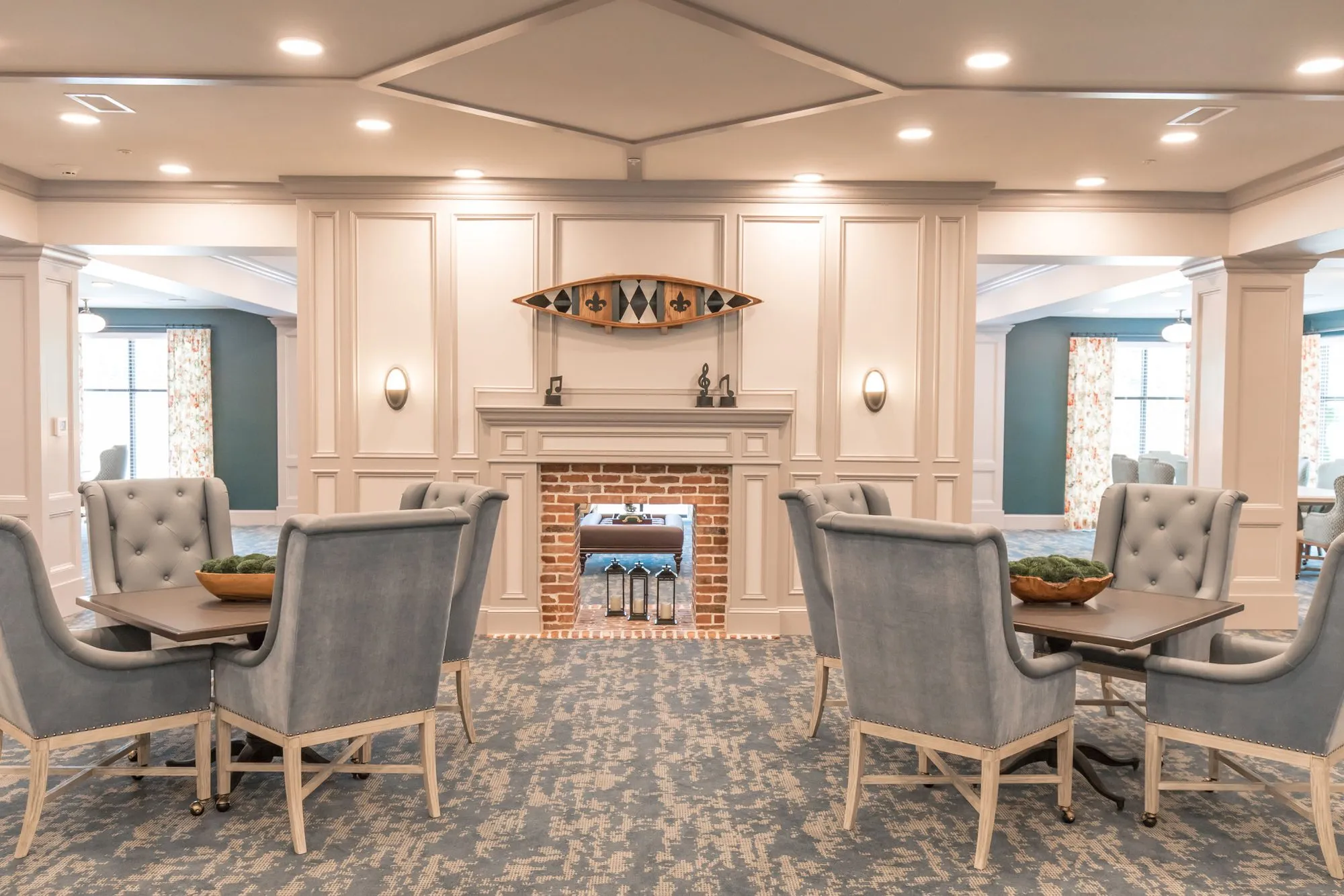 Senior living lounge with grey tufted wing chairs at game tables either side of a brick fireplace, a canoe hung above