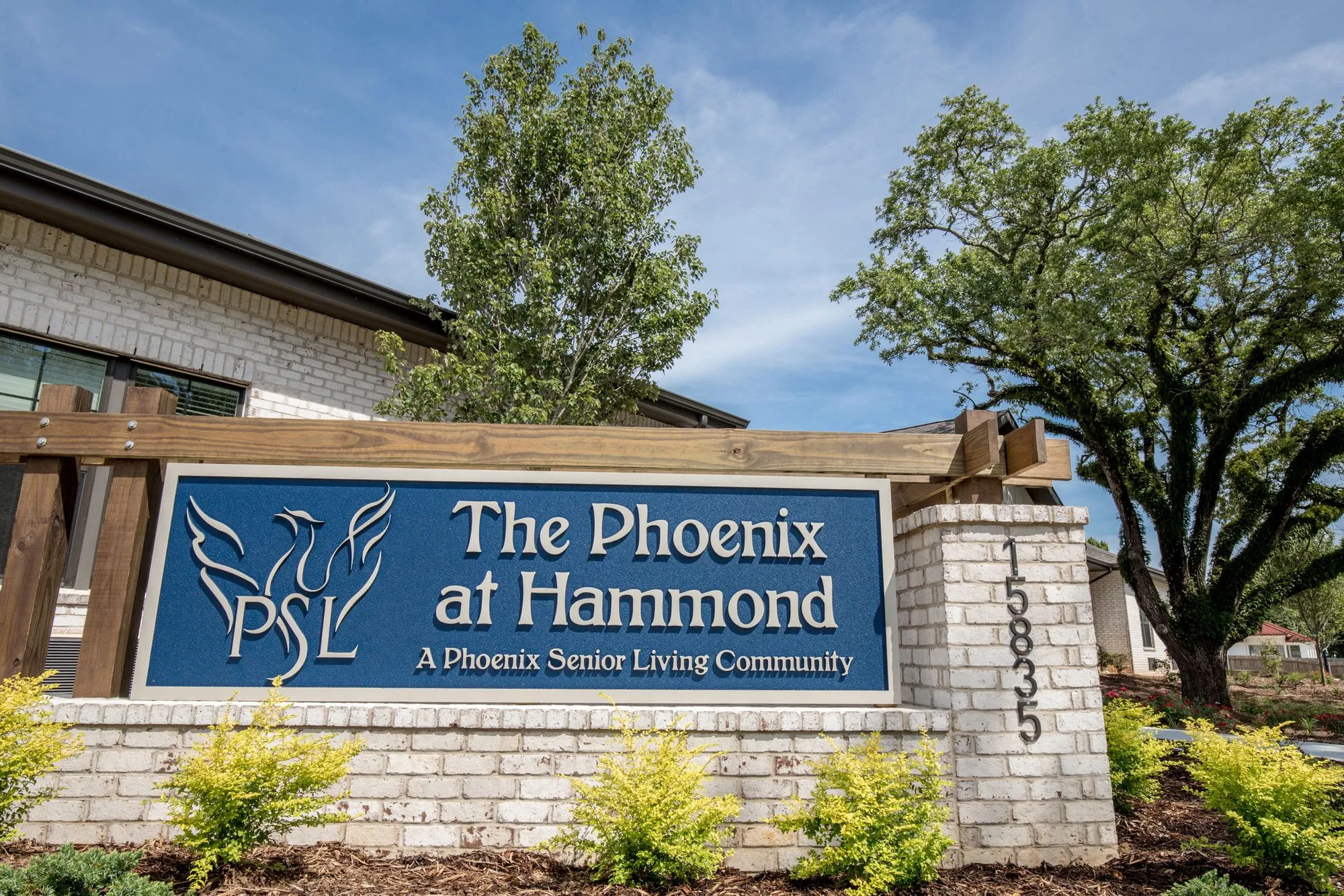 Senior living community exterior: brick and timber monument sign at the entrance, set in planted beds with oaks behind
