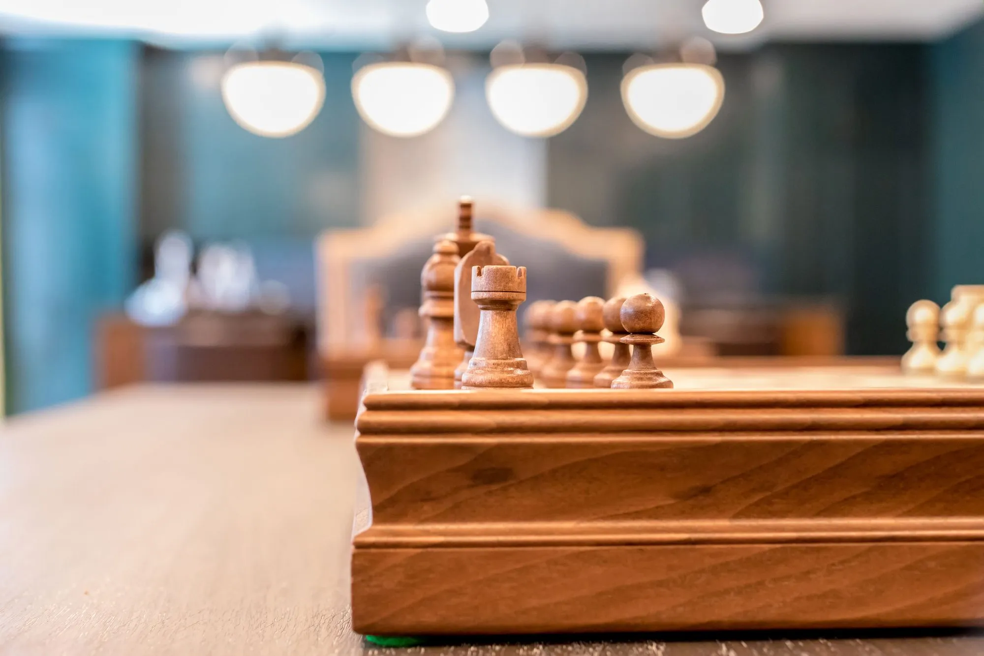 Senior living interior detail: wooden chess set on a table, globe pendants and teal walls blurred behind