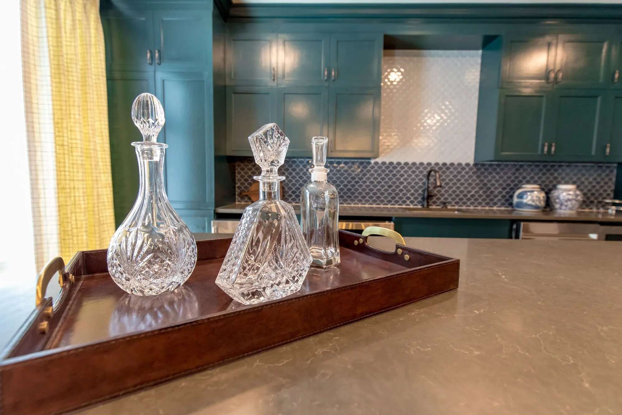 Senior living interior detail: cut glass decanters on a leather tray on a stone counter, dark teal cabinetry beyond