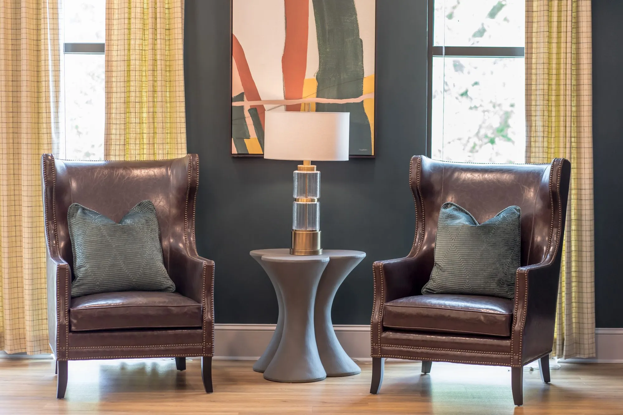 Senior living interior detail: pair of dark leather wing chairs with green pillows beside a lamp table, plaid curtains