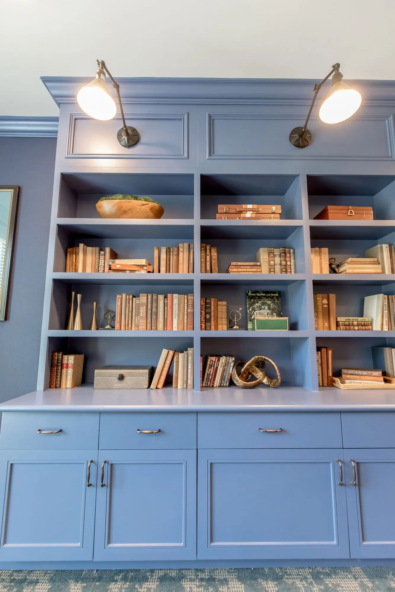 Senior living interior detail: periwinkle bookcase cabinetry with brass swing-arm lamps above shelves of books