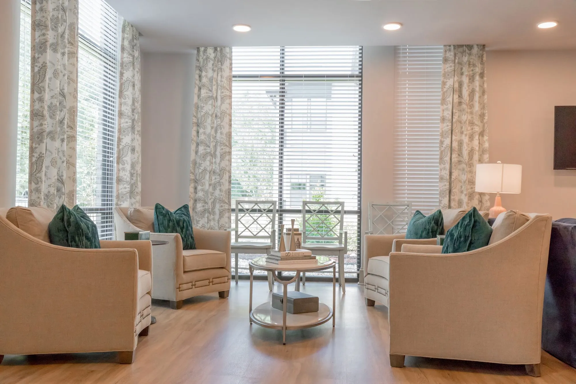 Senior living sitting area of linen armchairs with teal pillows around a round table by windows with botanical drapery