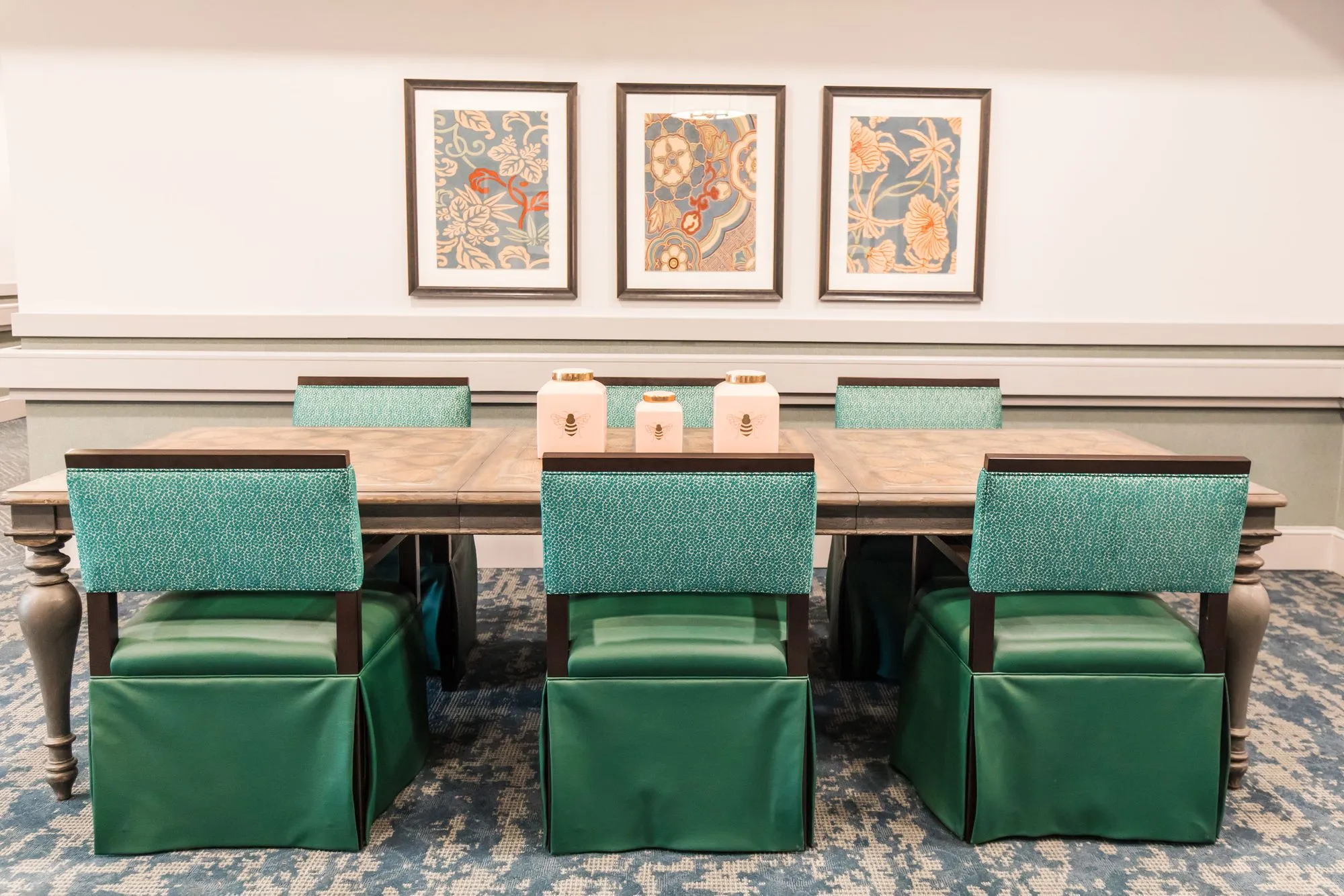 Senior living interior detail: long table with green skirted chairs with patterned backs beneath three framed floral prints