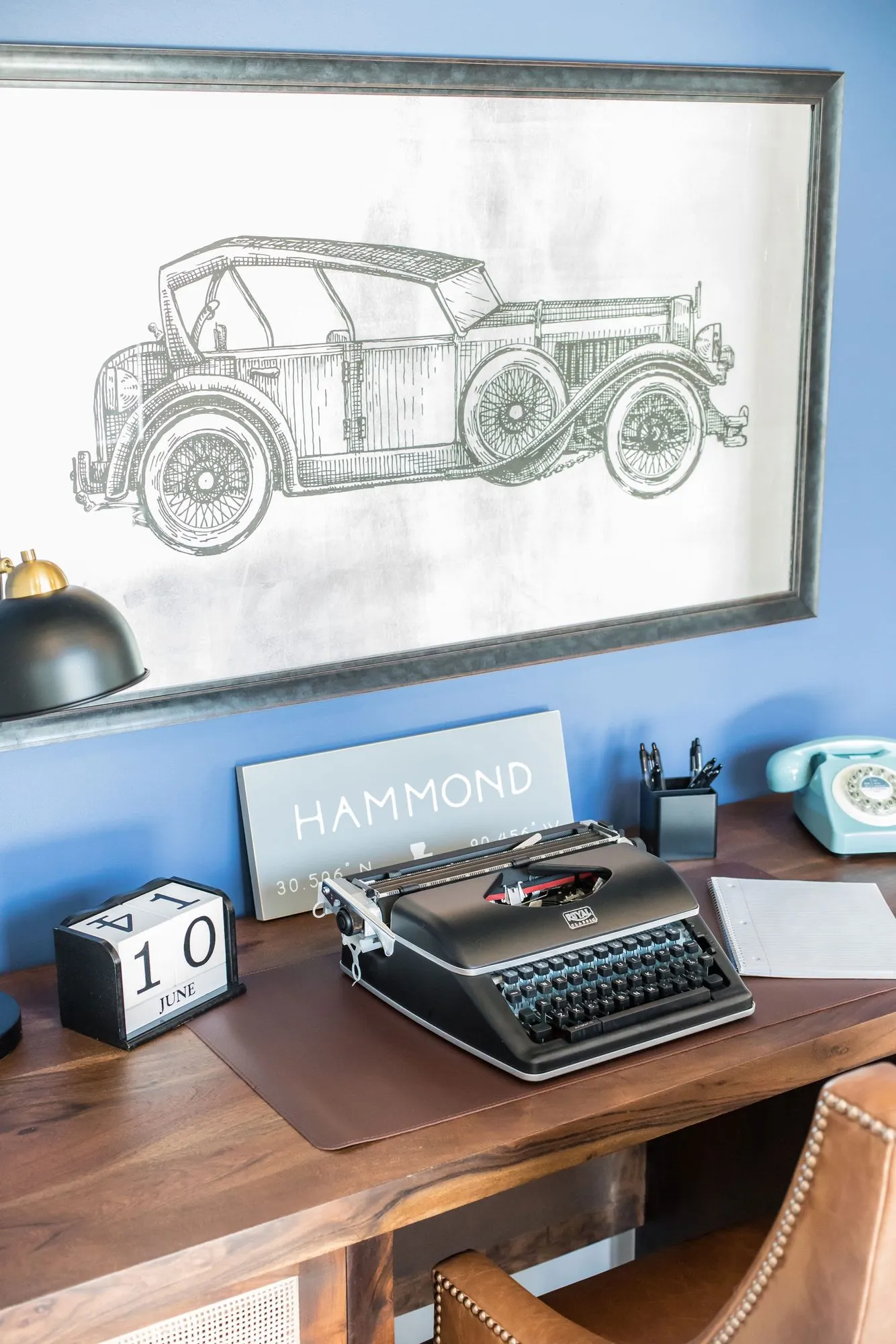 Senior living interior detail: black typewriter, flip calendar and teal phone on a wood desk under a vintage car drawing