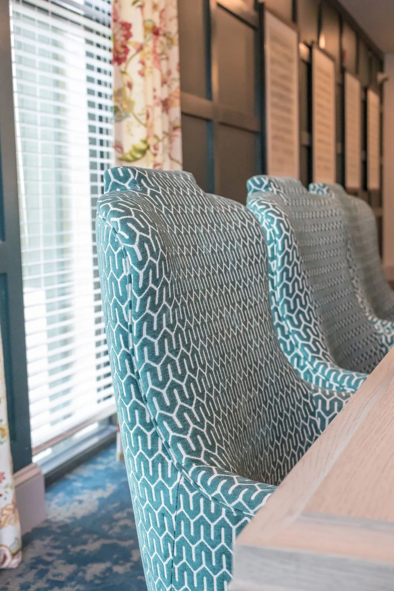 Senior living interior detail: teal geometric patterned high-back chairs lined up along a wood table by the window