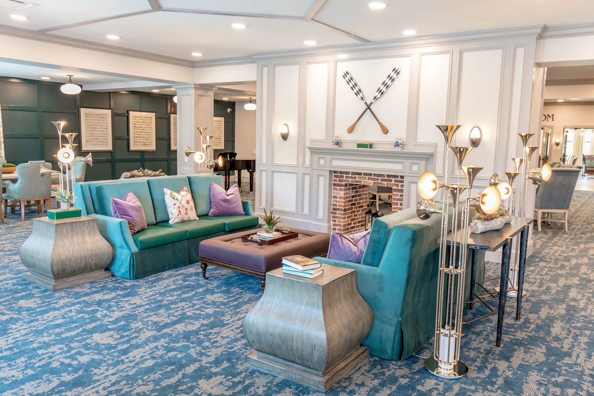 Senior living music room with teal sofas and lavender pillows around a purple ottoman by a brick fireplace under crossed oars