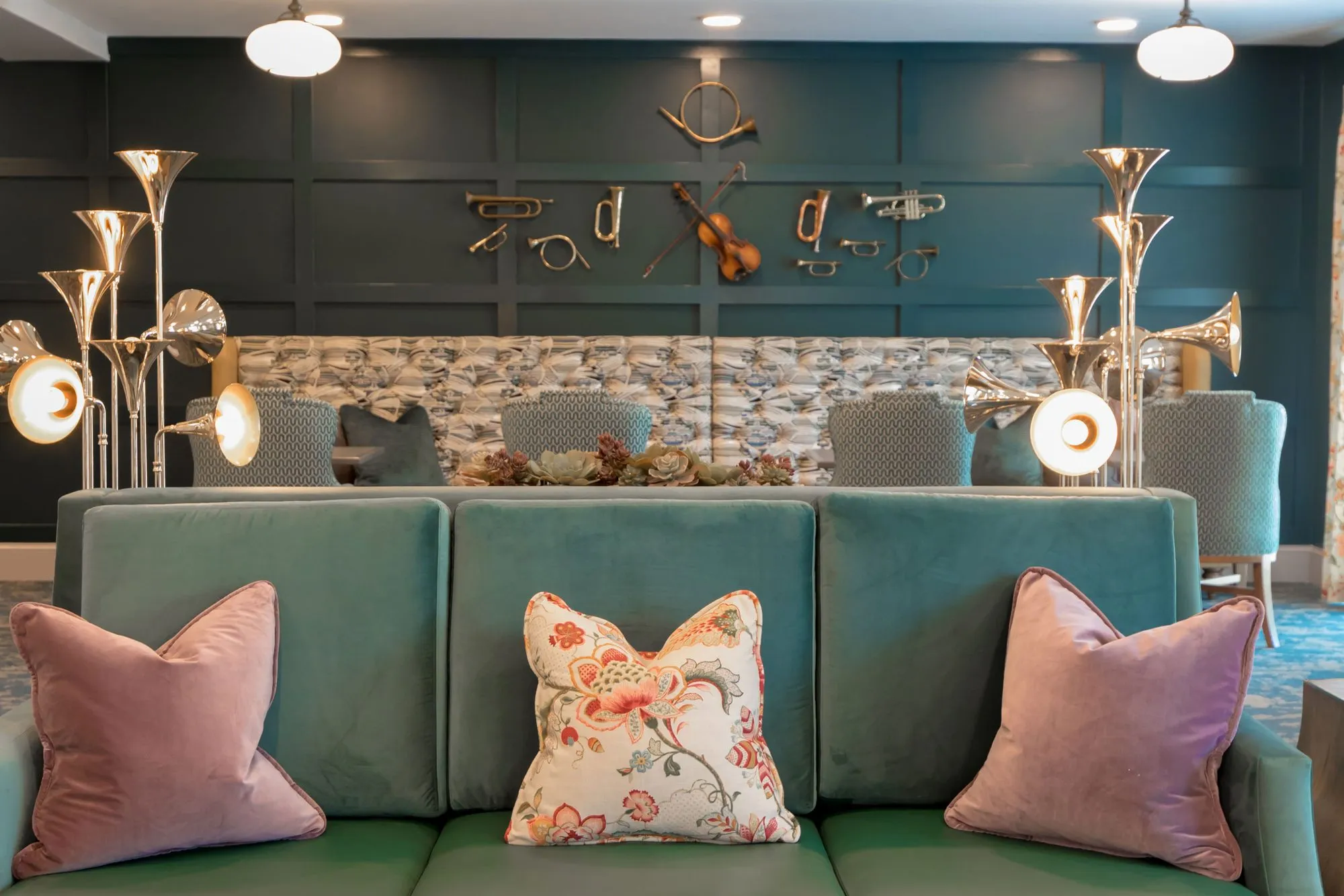 Senior living music room with a teal velvet banquette and floral pillows between horn floor lamps, brass instruments behind