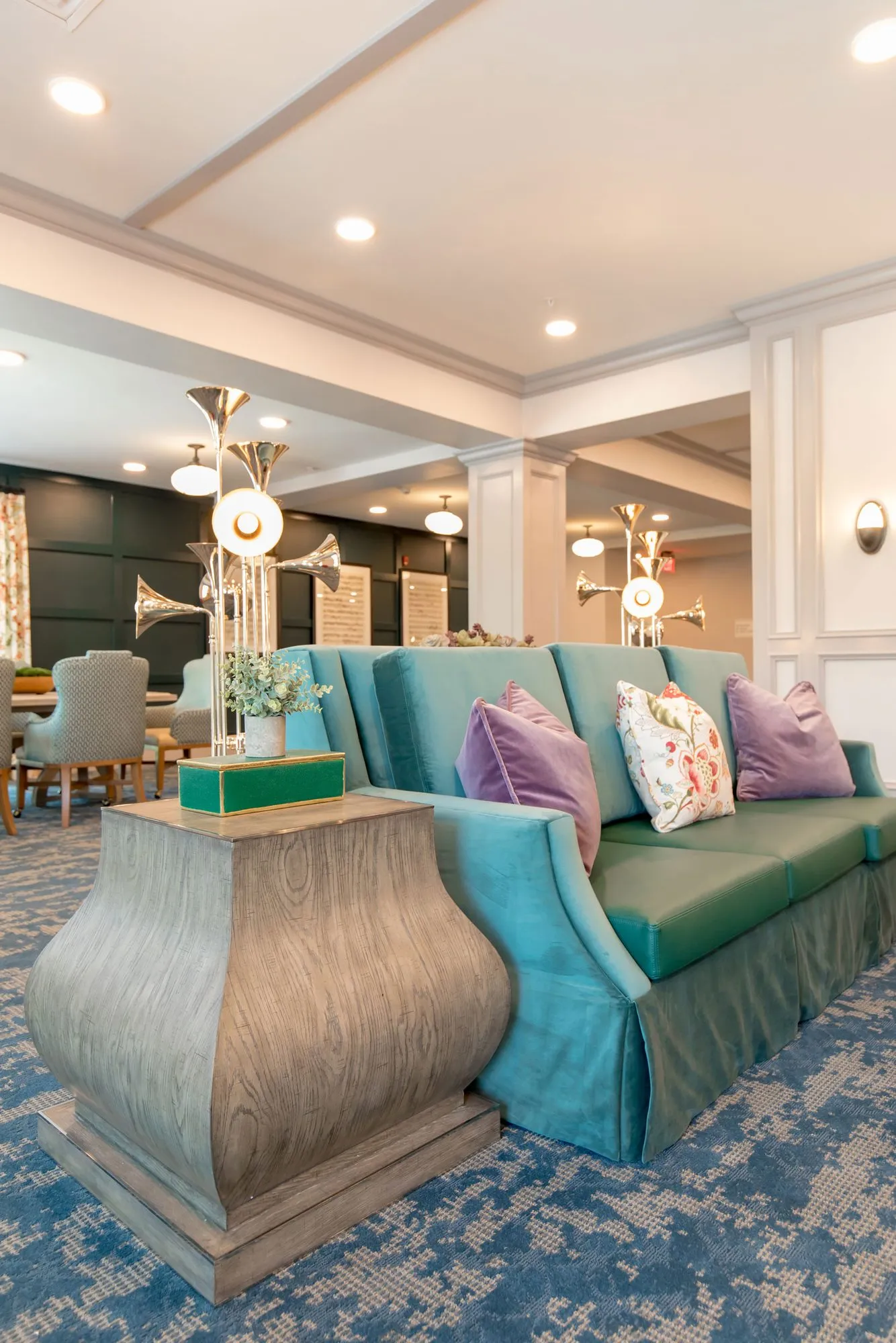 Senior living music room with a teal skirted sofa and lavender pillows beside a silver wood chest, horn lamps behind