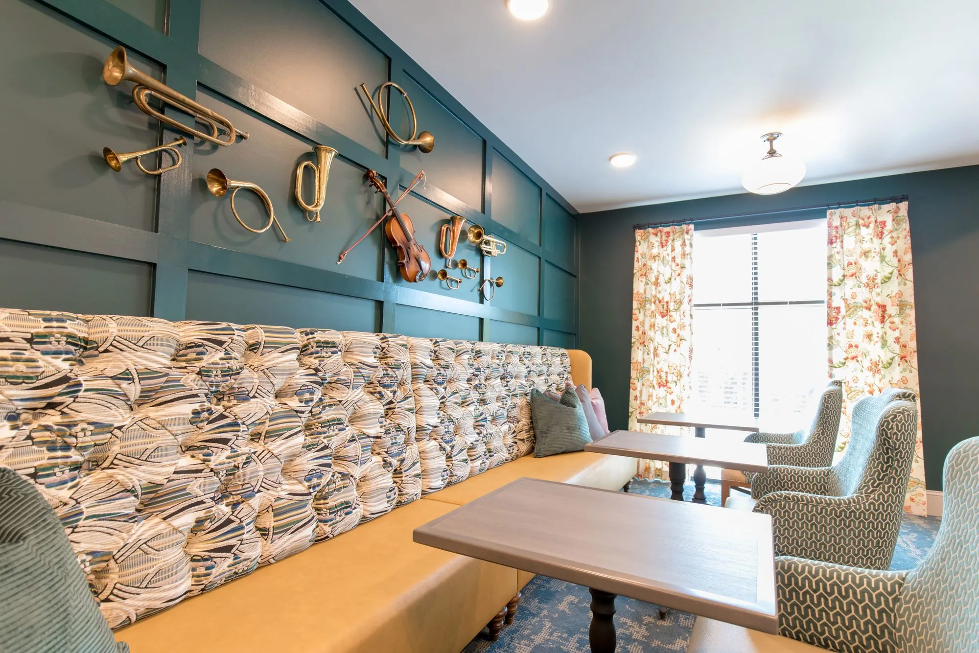 Senior living bistro with a patterned banquette and tables below brass instruments on teal panelling, floral curtains beyond