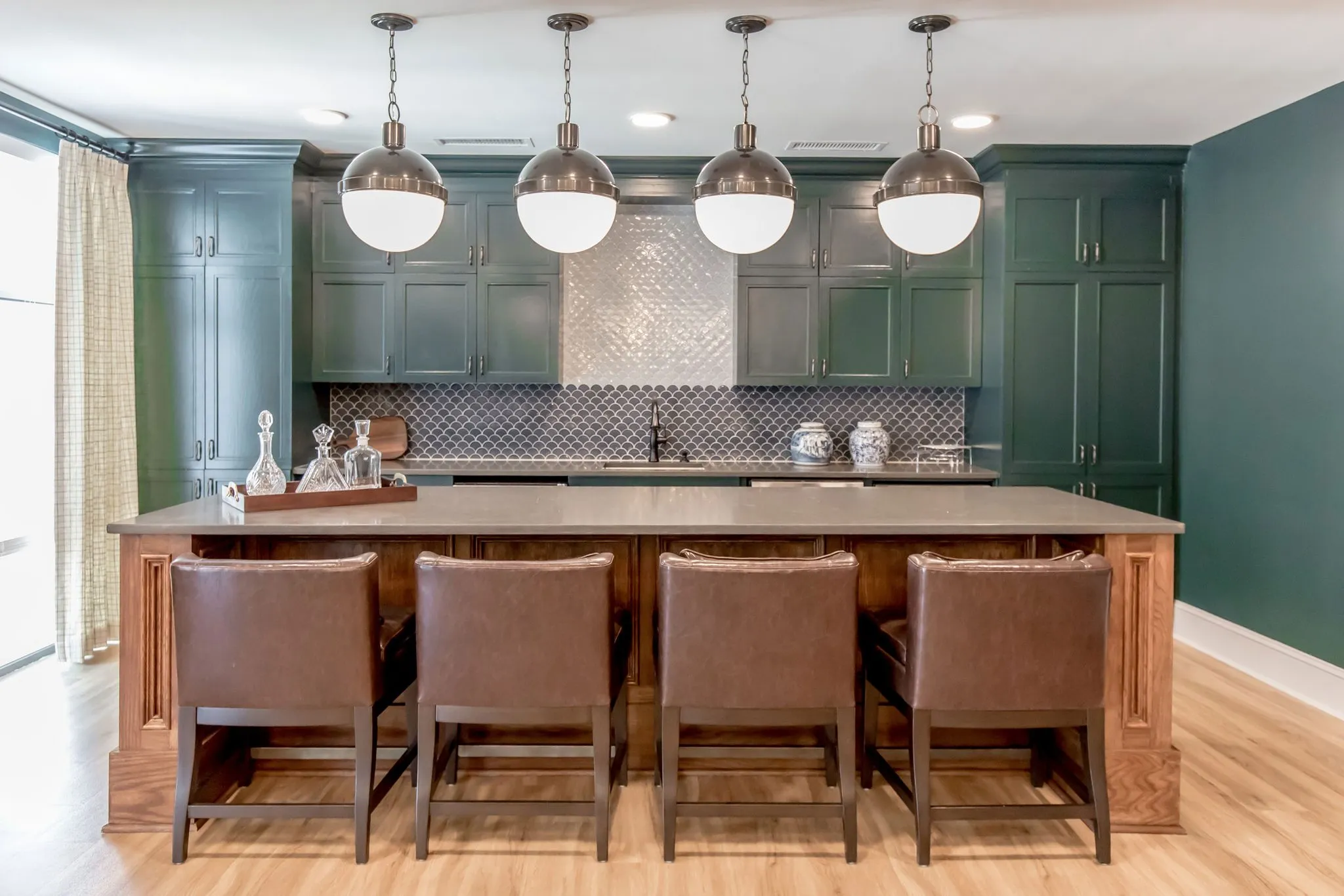 Senior living demonstration kitchen with dark green cabinetry and a silver fish-scale backsplash behind leather stools