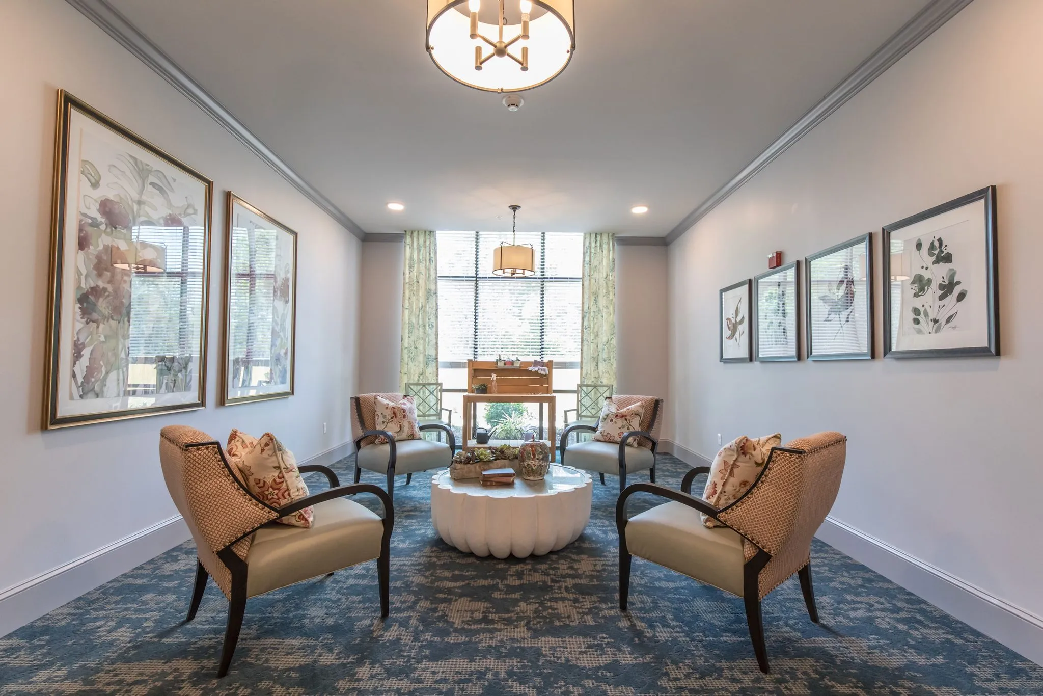 Senior living sitting room with patterned armchairs drawn around a scalloped white table below botanical prints