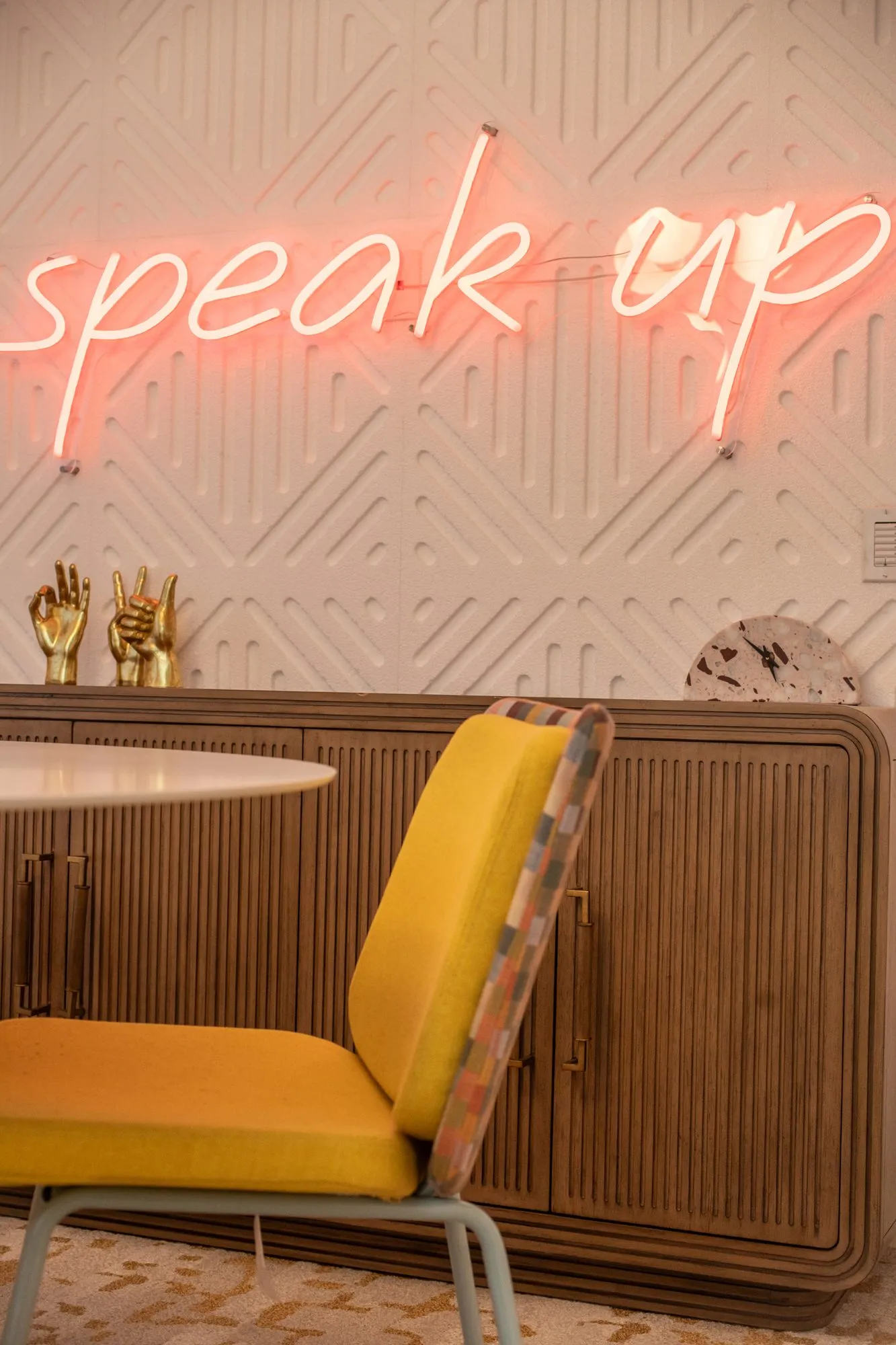 Student housing interior detail: pink neon reading speak up on white relief panels above a fluted sideboard, a yellow chair