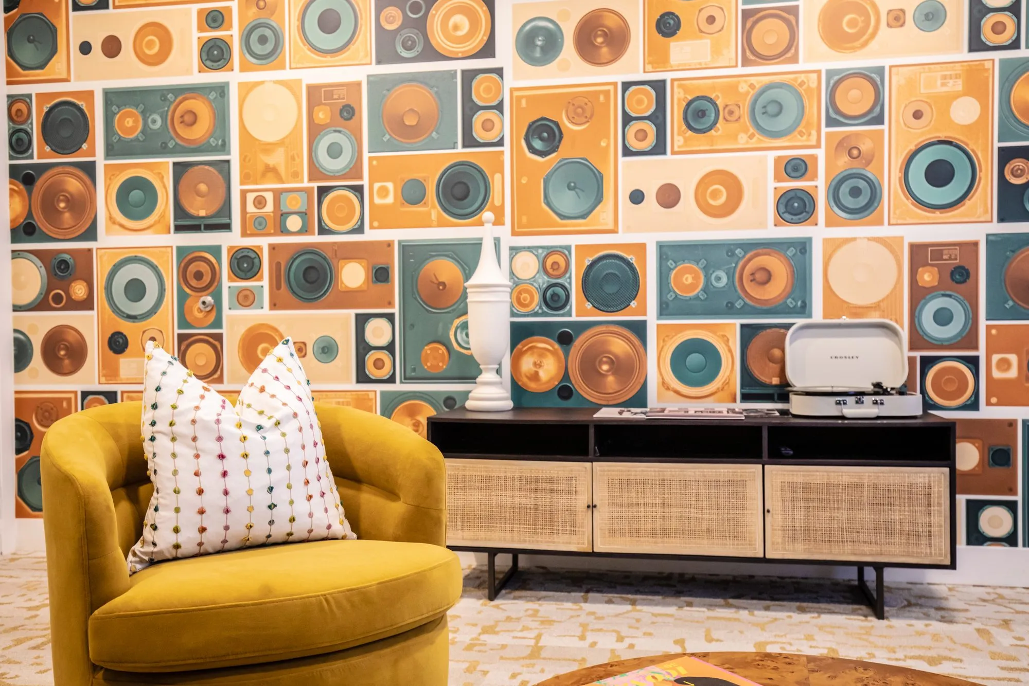Student housing interior detail: mustard swivel chair, rattan media console and record player against speaker print wallpaper