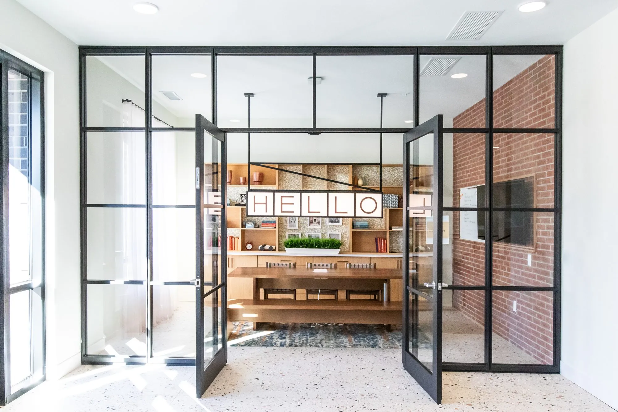 Student housing reception through open black steel framed glass doors, a HELLO letter sign on wood shelving by brick