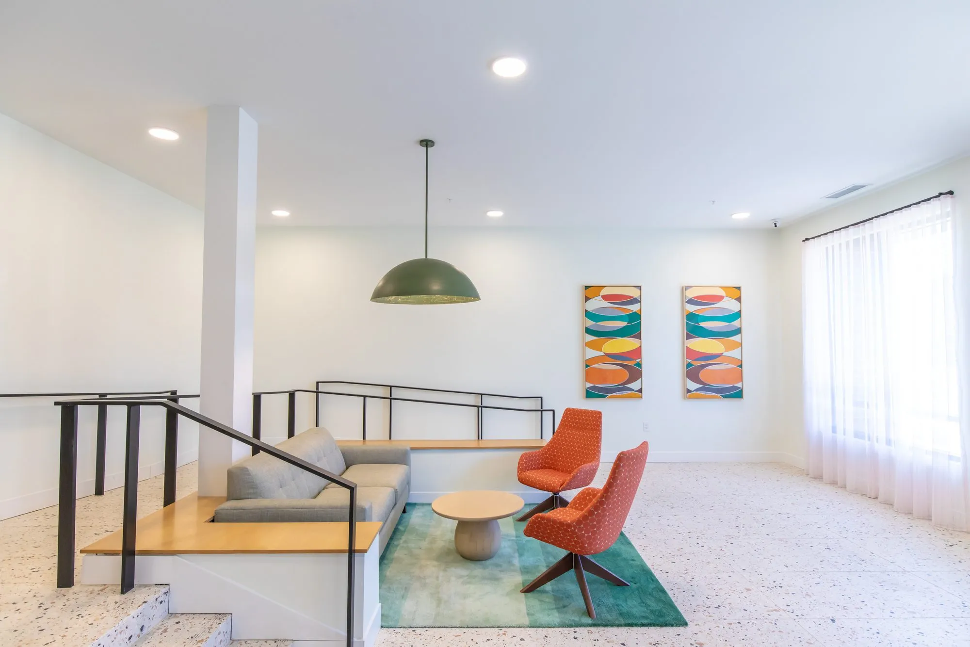 Student housing amenity sunken lounge with a grey built-in sofa and orange chairs on a teal rug under a green dome pendant