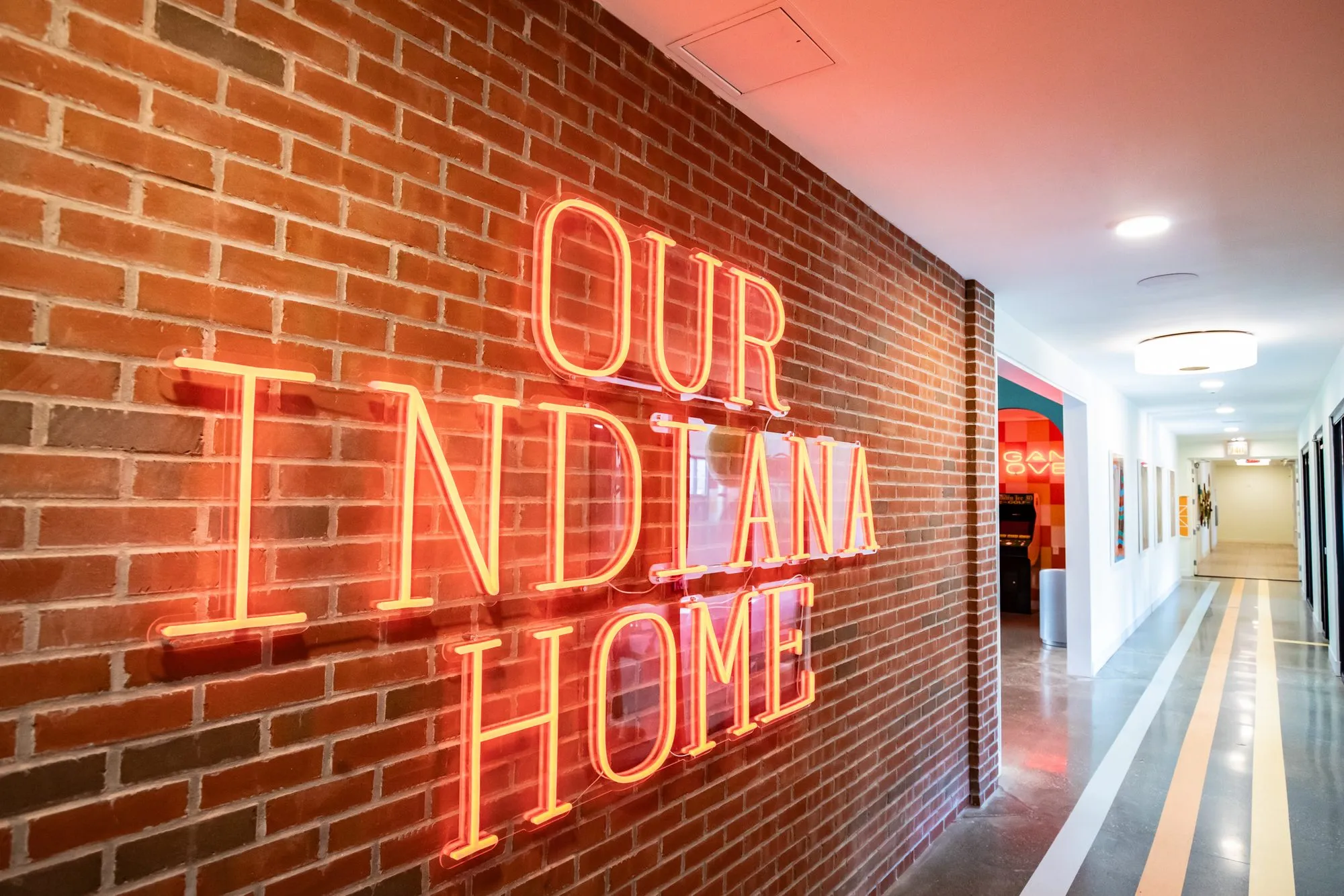 Student housing amenity corridor: orange neon reading Our Indiana Home on brick, seen at an angle over the striped floor
