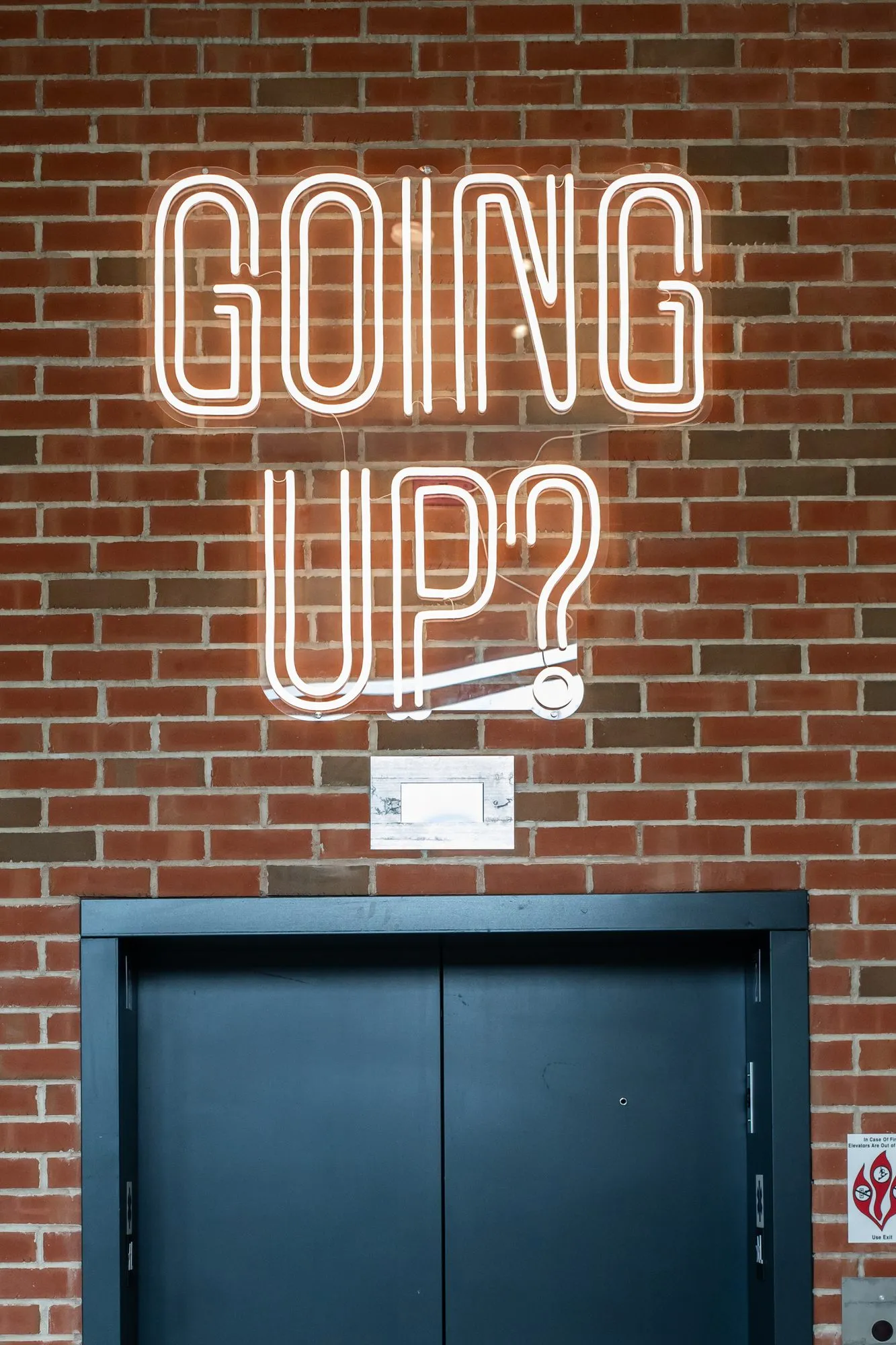 Student housing interior detail: white neon reading Going Up? on red brick above the elevator doors