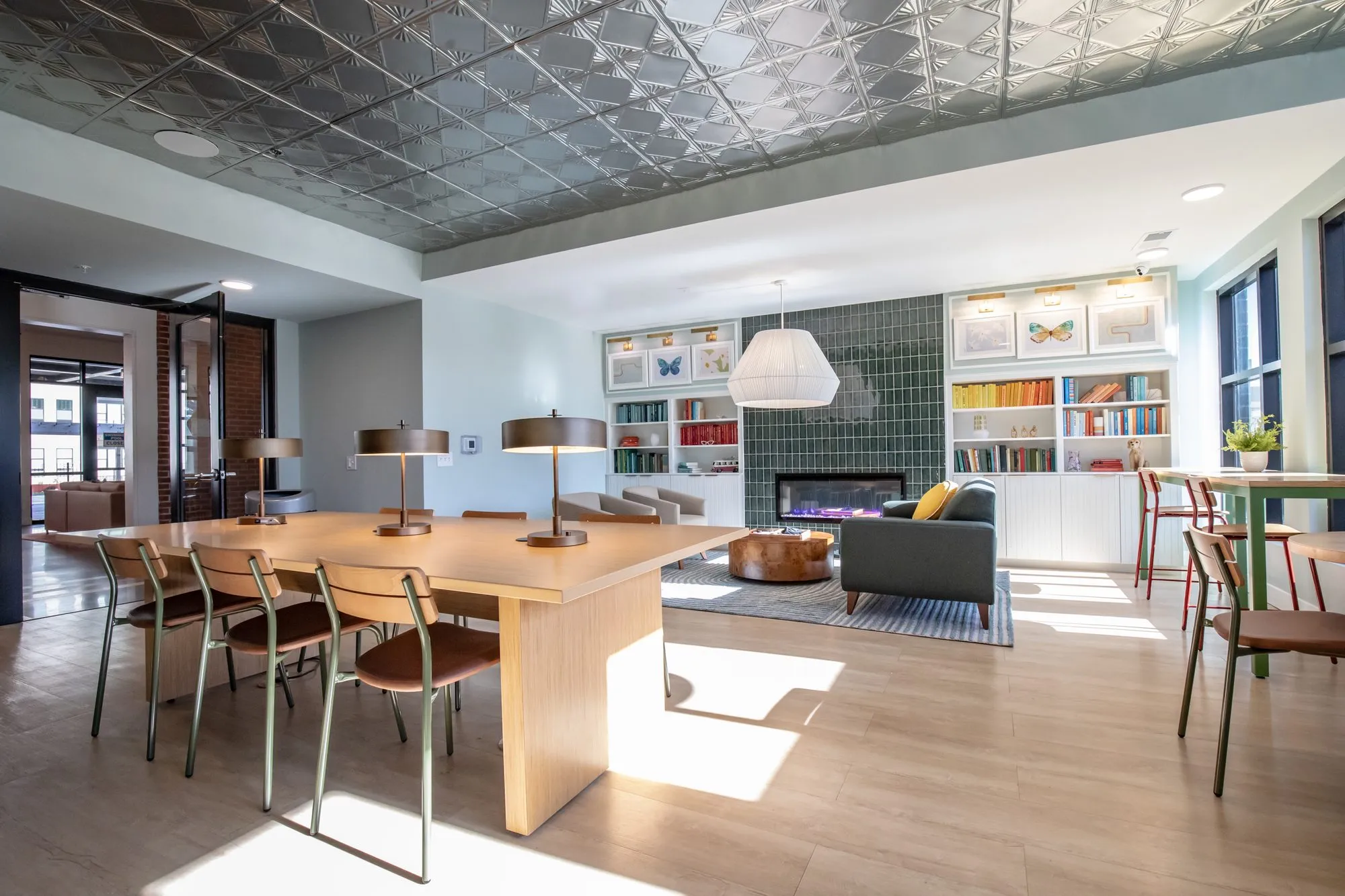 Student housing amenity study lounge with a long wood table and brass lamps under a pressed tin ceiling, a tiled fireplace