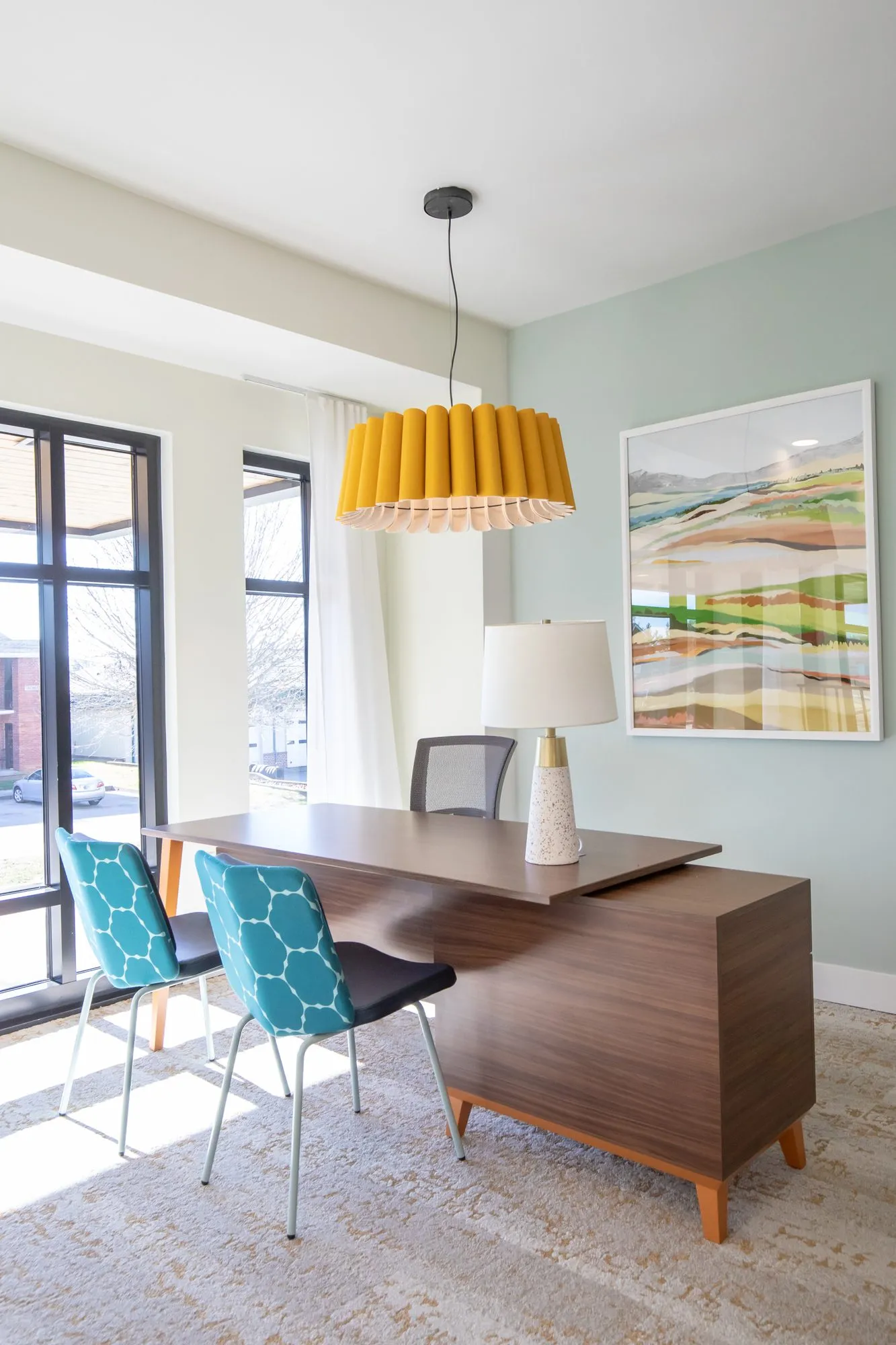 Student housing amenity office with a walnut desk, teal patterned chairs and a yellow pleated pendant beside tall windows