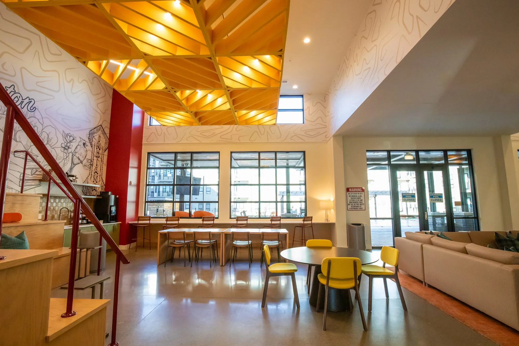 Student housing amenity double-height lounge under an orange timber lattice ceiling, red stair rail and yellow chairs
