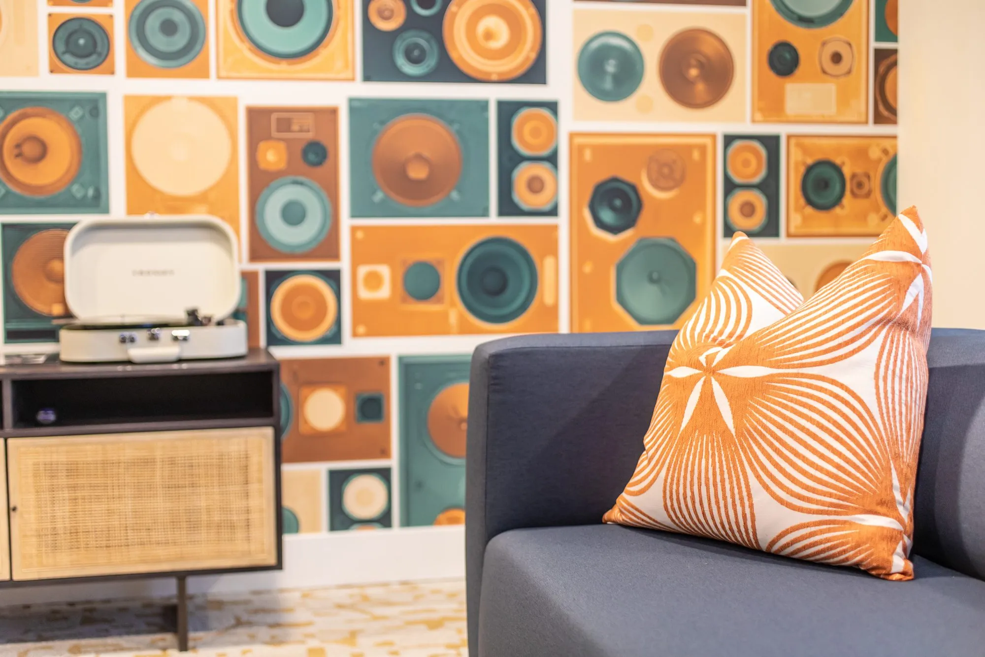 Student housing interior detail: grey sofa with an orange pillow beside a rattan console and record player, speaker wallpaper