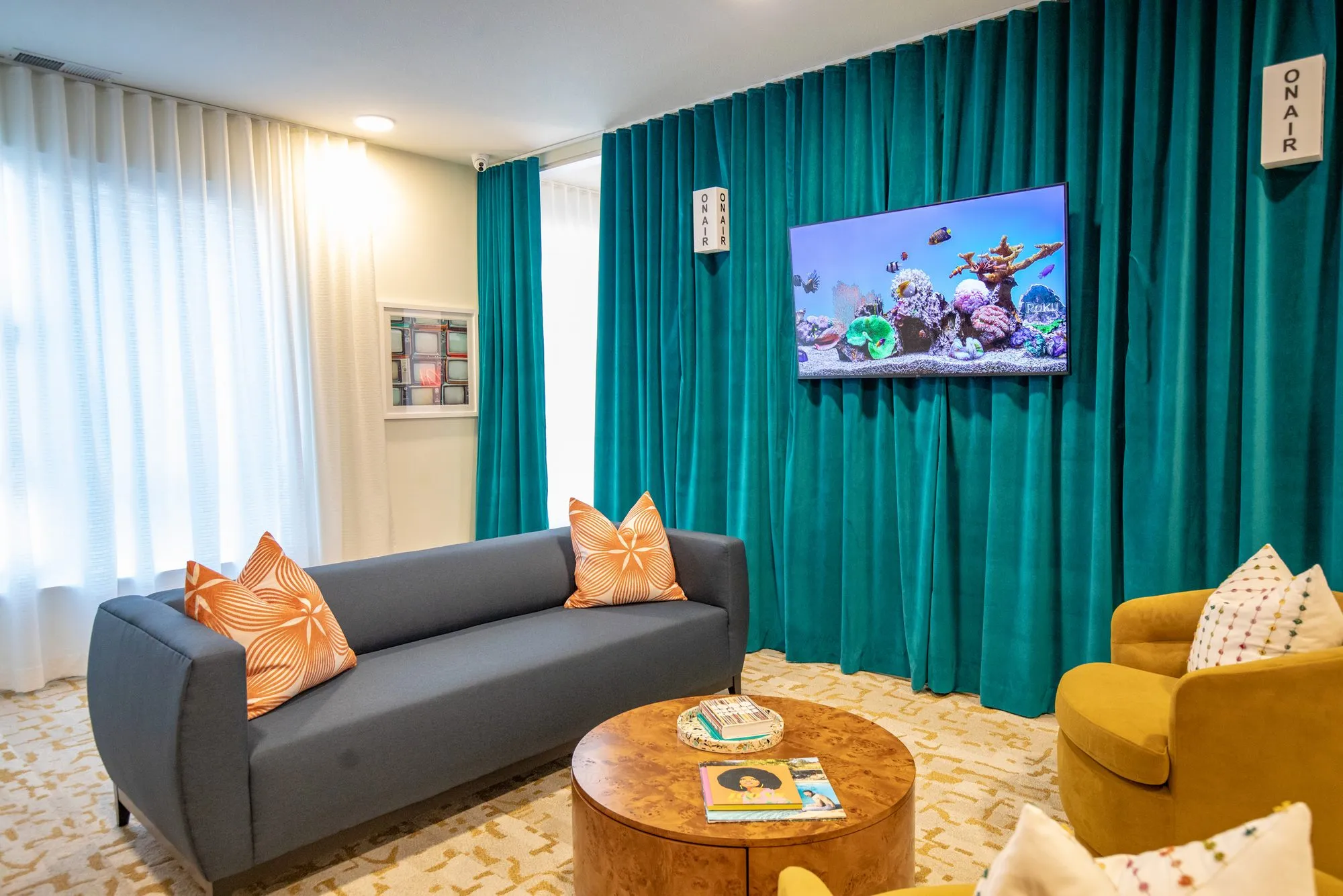 Student housing amenity lounge with a grey sofa, mustard armchair and burl coffee table before teal curtains and a screen