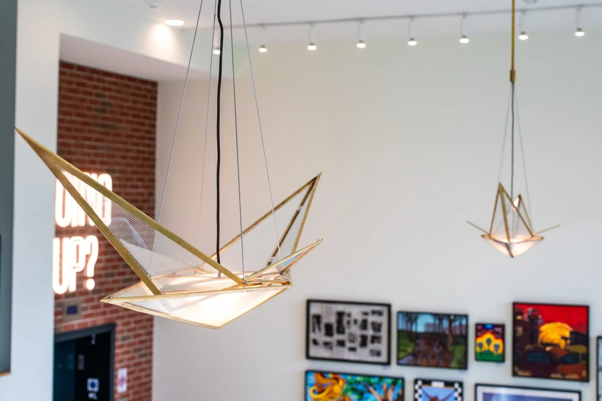 Student housing interior detail: brass geometric pendants before a gallery wall of framed art and a neon sign on brick