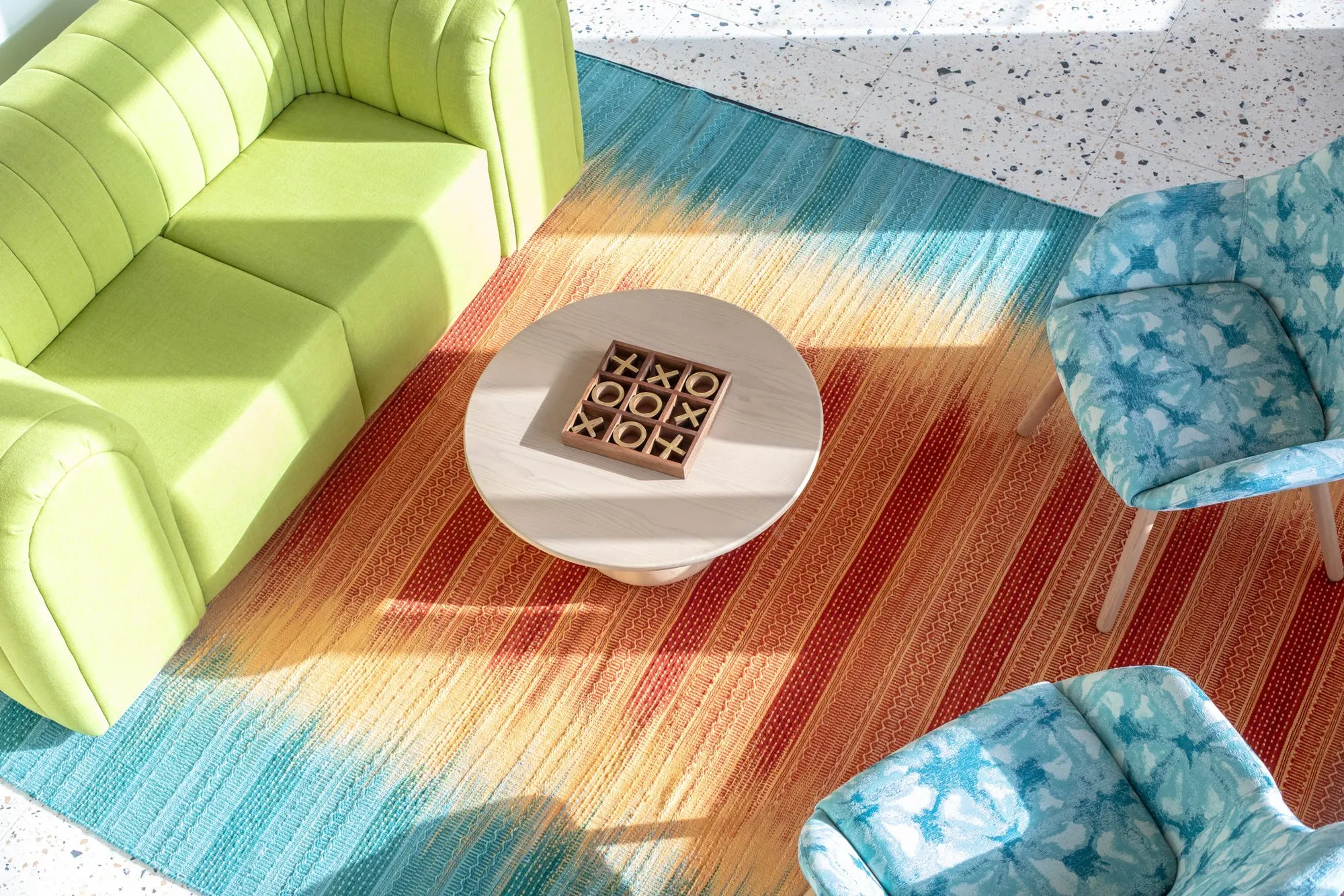 Student housing amenity lounge from above: a lime green sofa, blue floral chairs and a tic tac toe table on a striped rug