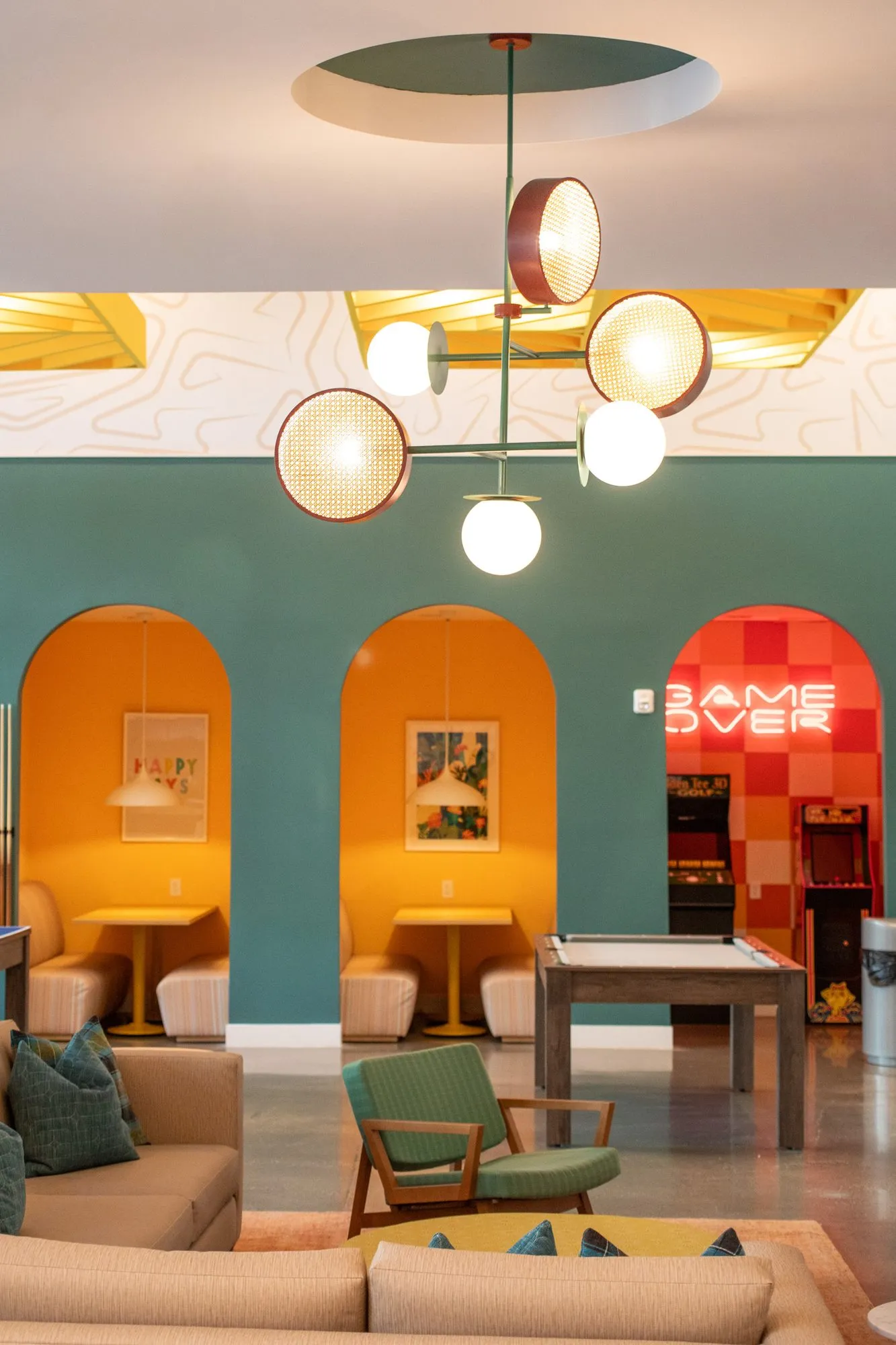 Student housing amenity game room past a sofa and green armchair to orange arched booths, a globe chandelier overhead