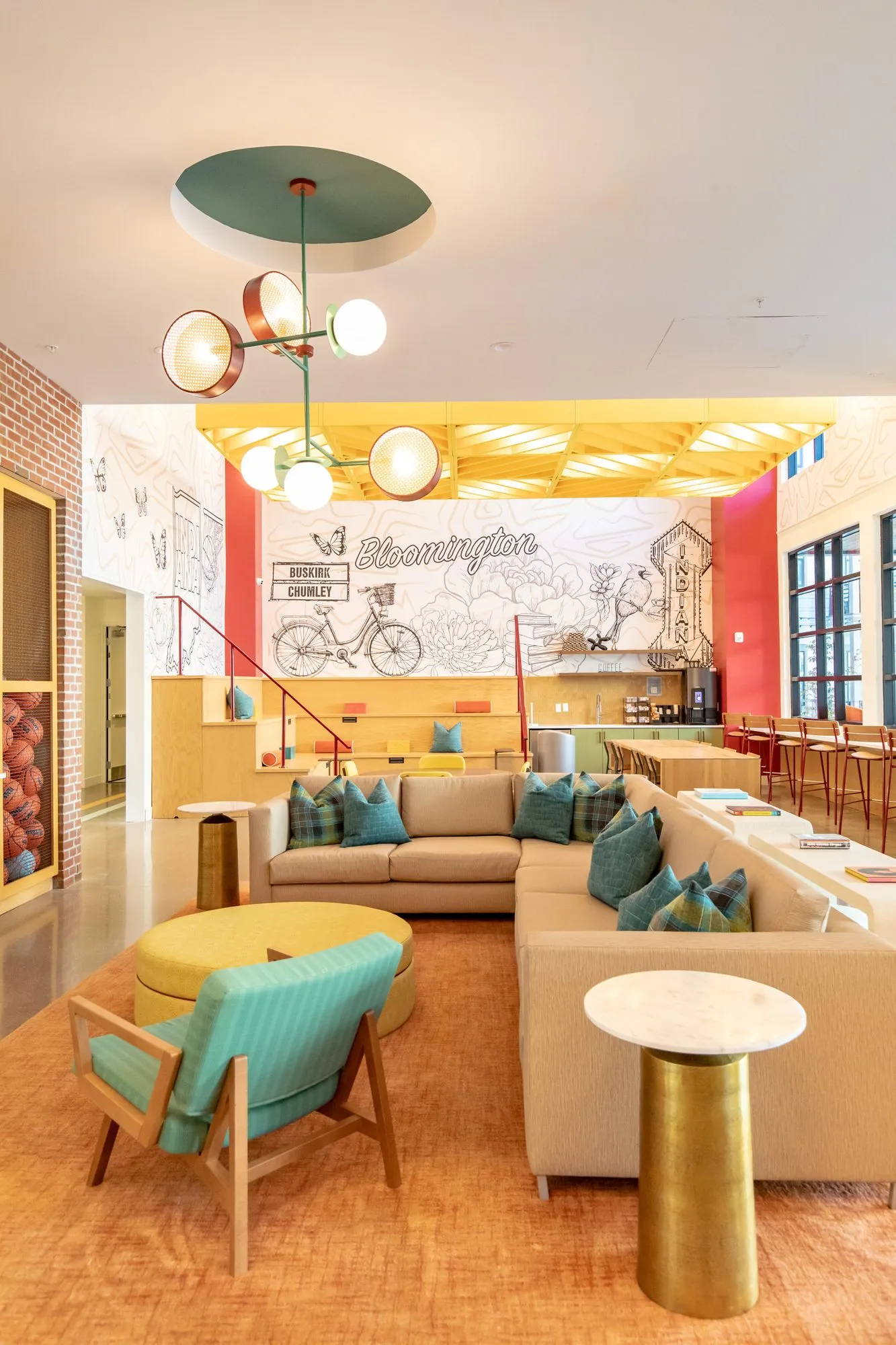 Student housing amenity lounge with a sectional and teal armchair under a globe chandelier, the bicycle mural beyond