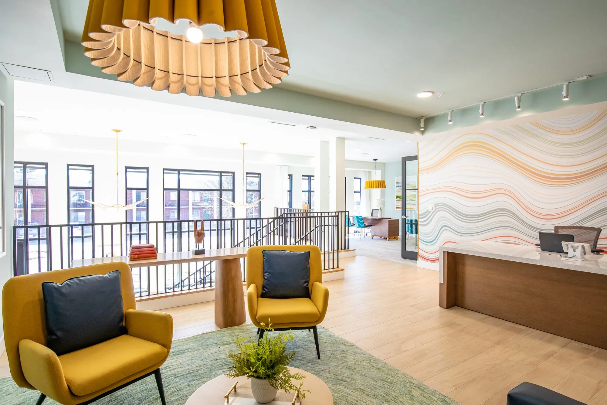 Student housing amenity lobby with mustard armchairs on a green rug under a pleated pendant, reception desk and wave mural