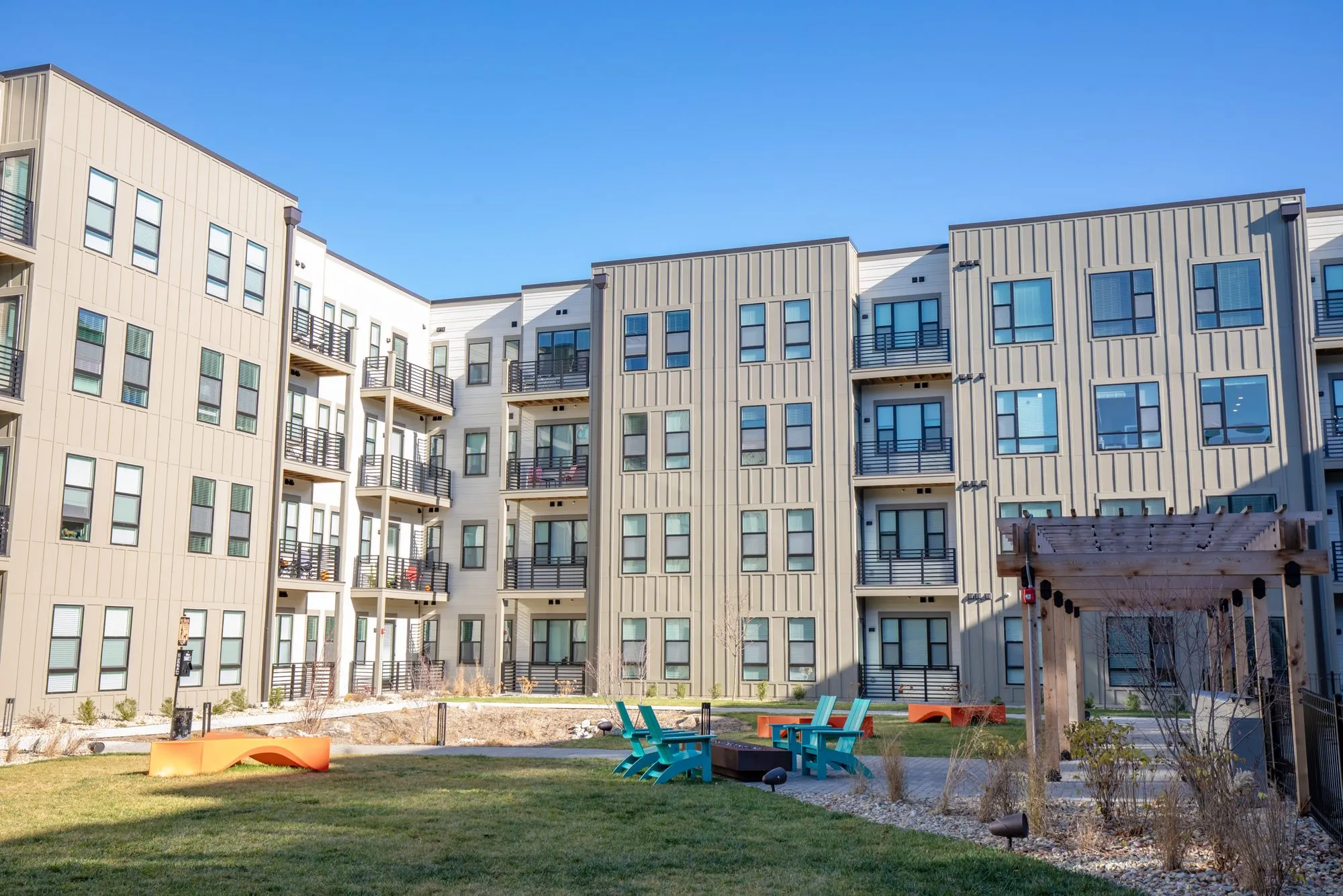 Student housing courtyard lawn with teal Adirondack chairs around a fire pit and a pergola before grey panelled wings