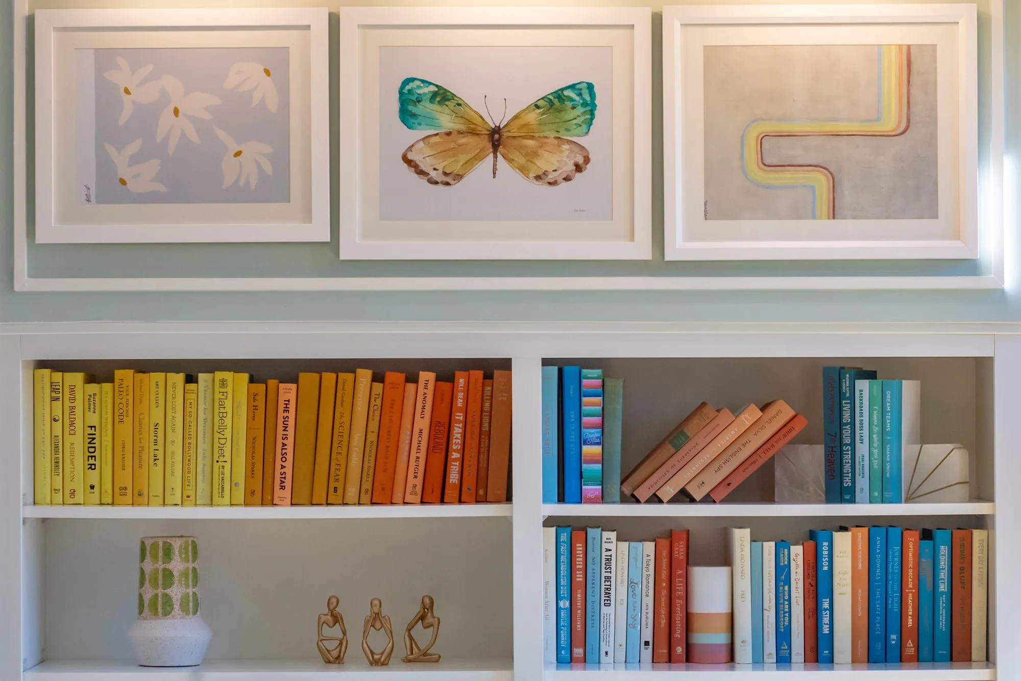 Student housing interior detail: colour-sorted books on white shelving below framed flower, butterfly and rainbow prints
