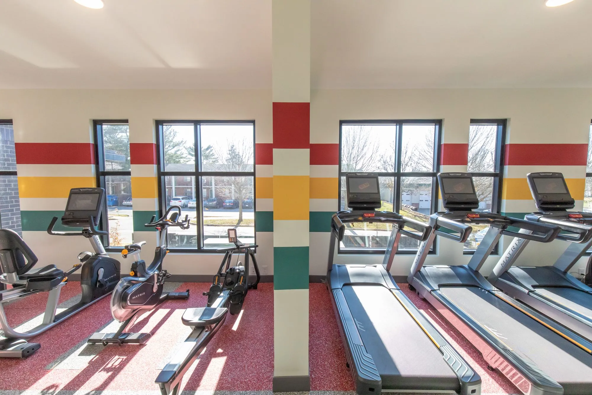 Student housing amenity fitness centre with treadmills and ellipticals on red rubber flooring, windows banded in colour