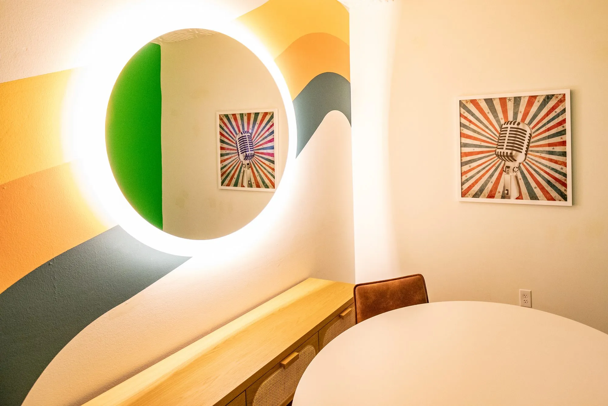 Student housing interior: backlit round mirror on a curved striped wall mural, microphone prints and a round white table