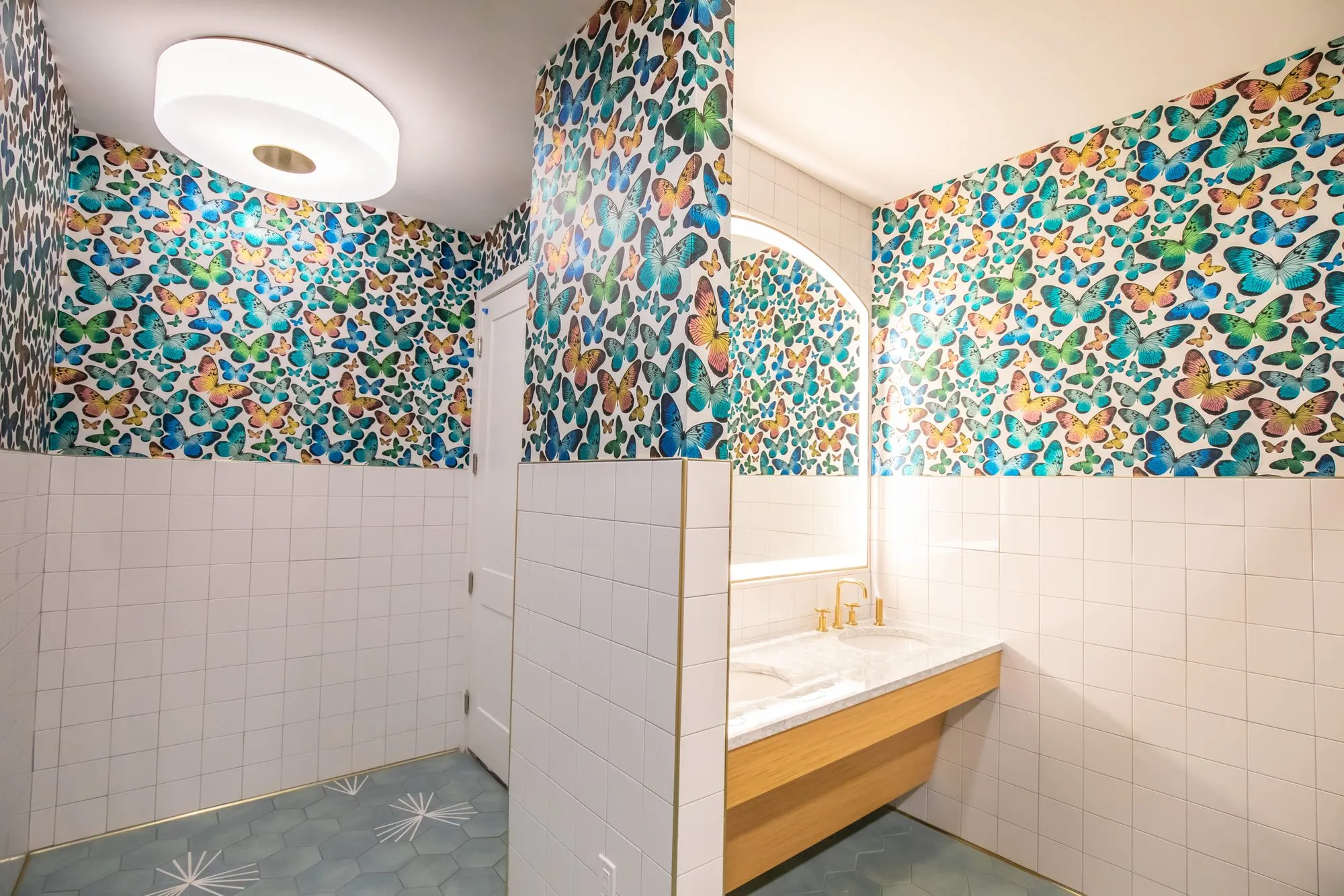 Student housing amenity restroom with butterfly wallpaper above white tile, a wood vanity with brass taps and arched mirror