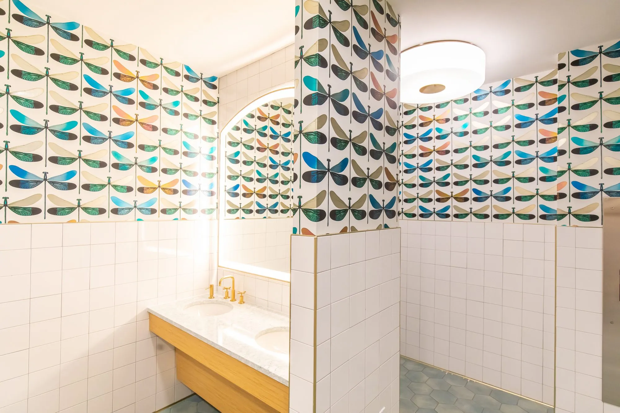 Student housing amenity restroom with dragonfly wallpaper above white tile, a marble vanity with brass taps, arched mirror