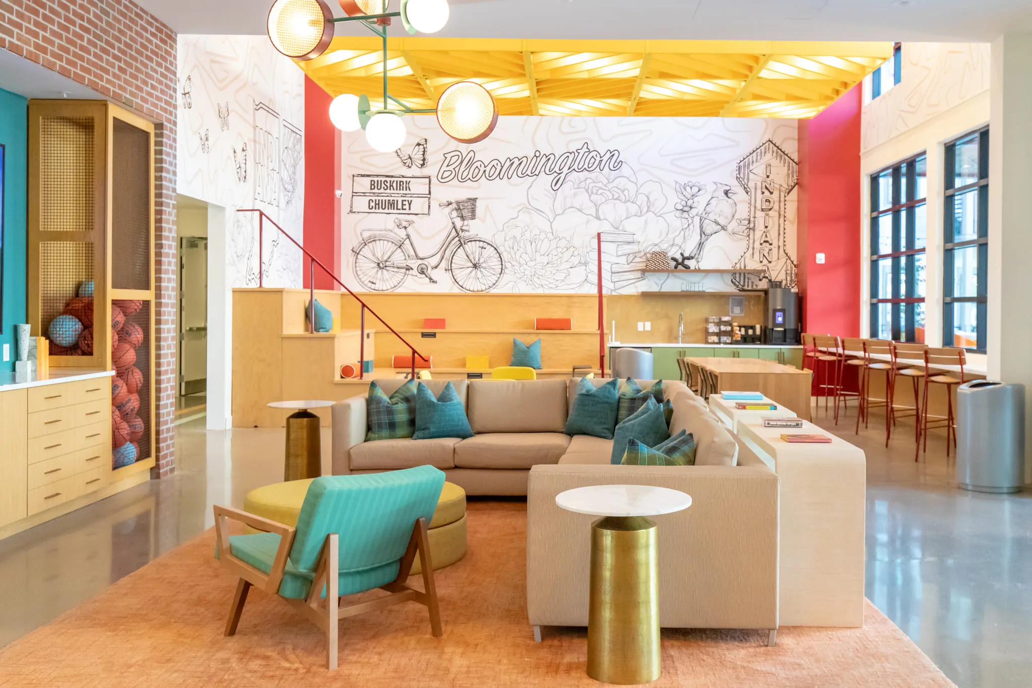 Student housing amenity lounge with a beige sectional, teal pillows and a teal armchair before a line drawn bicycle mural