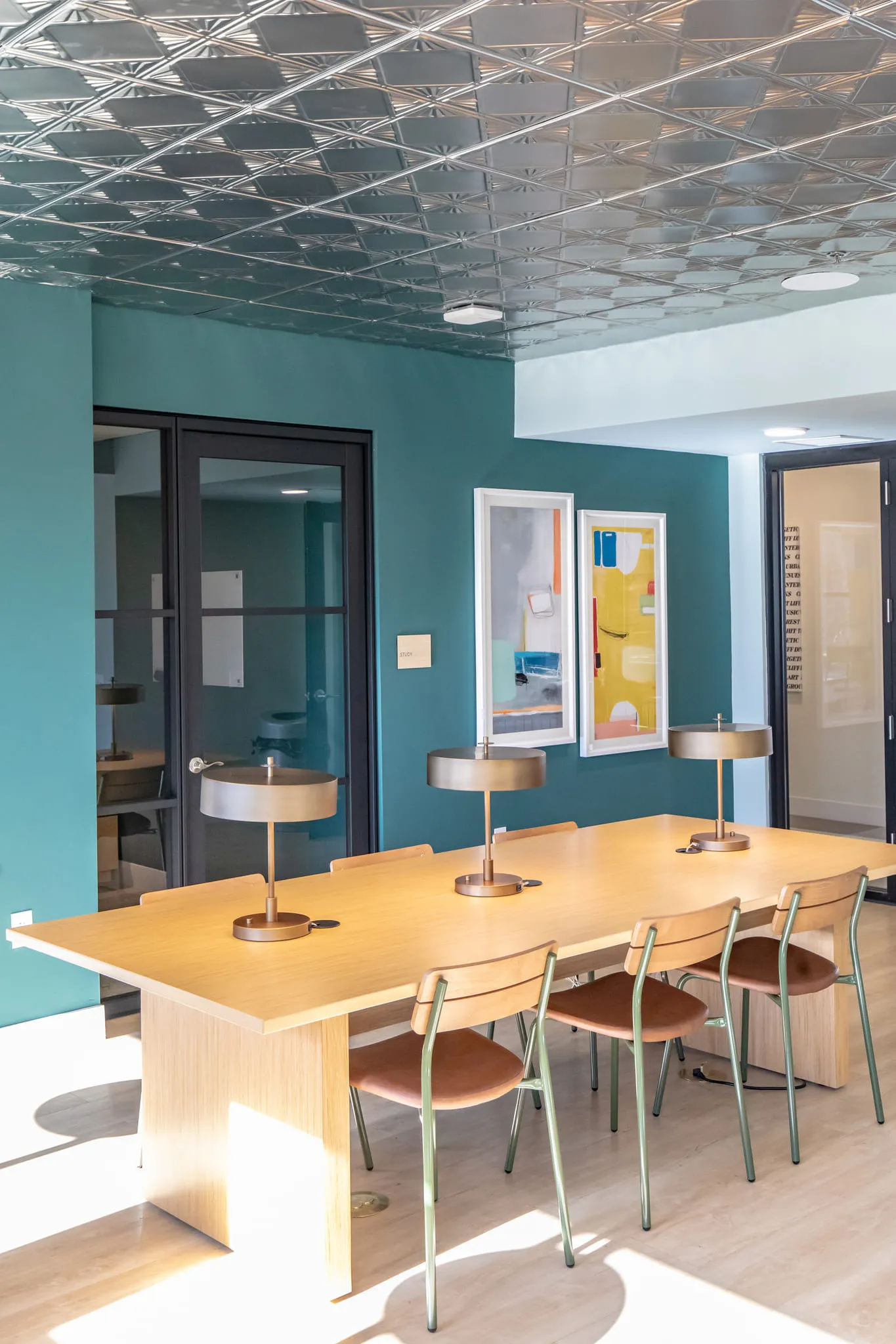 Student housing amenity study lounge with a long wood table and brass lamps under a pressed tin ceiling, prints on teal