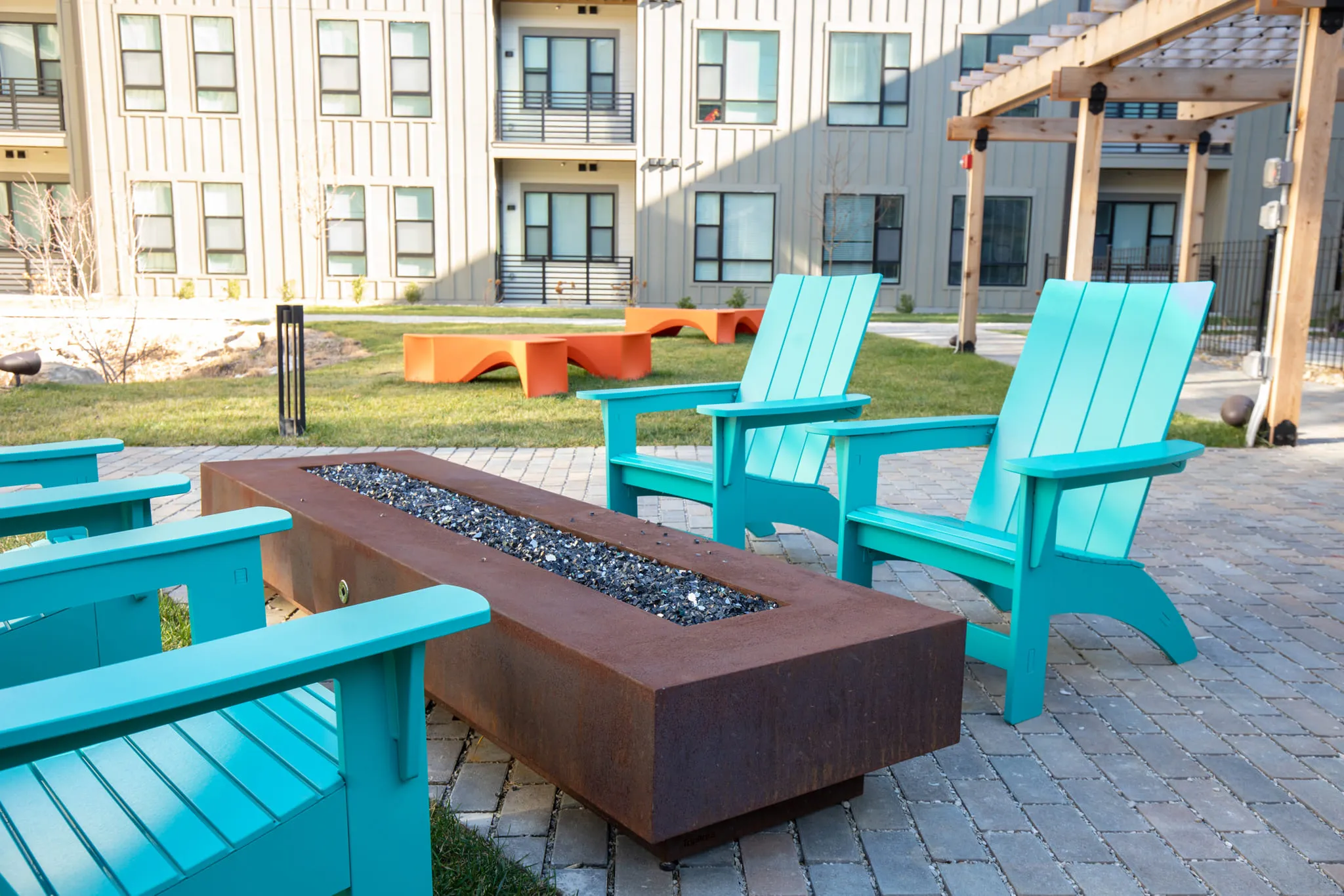 Student housing courtyard with turquoise Adirondack chairs around a long gas fire table on the paved terrace